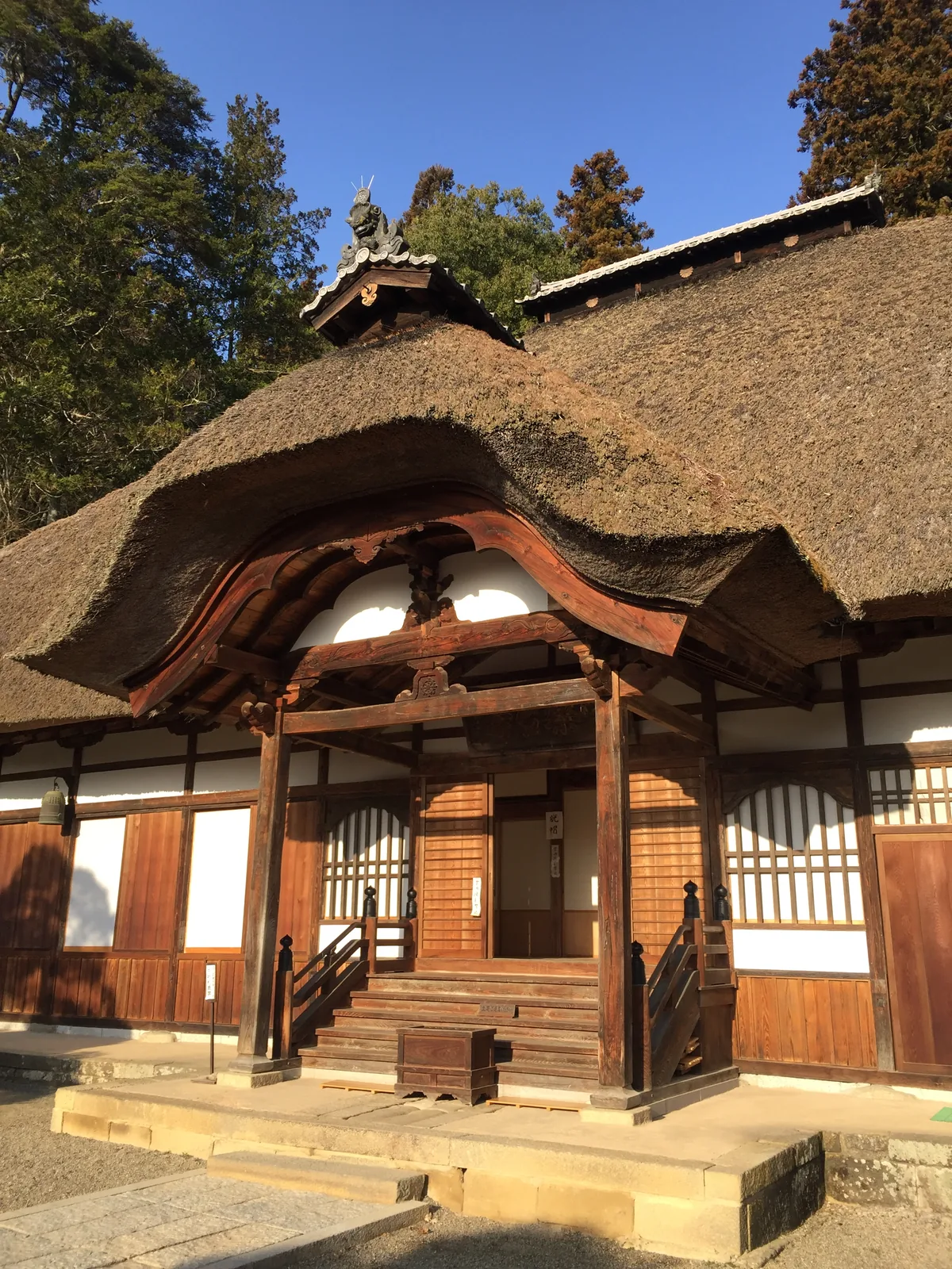 常樂寺本堂的向拜（こうはい，正面伸出的禮拜前廊）近景：茅的下襬自己彎成唐破風的弧，向拜柱是角柱，中備裝著雲形的蟇股（かえるまた，梁上的蛙腿形構件），左右各一對花頭窓，左端吊著一口半鐘