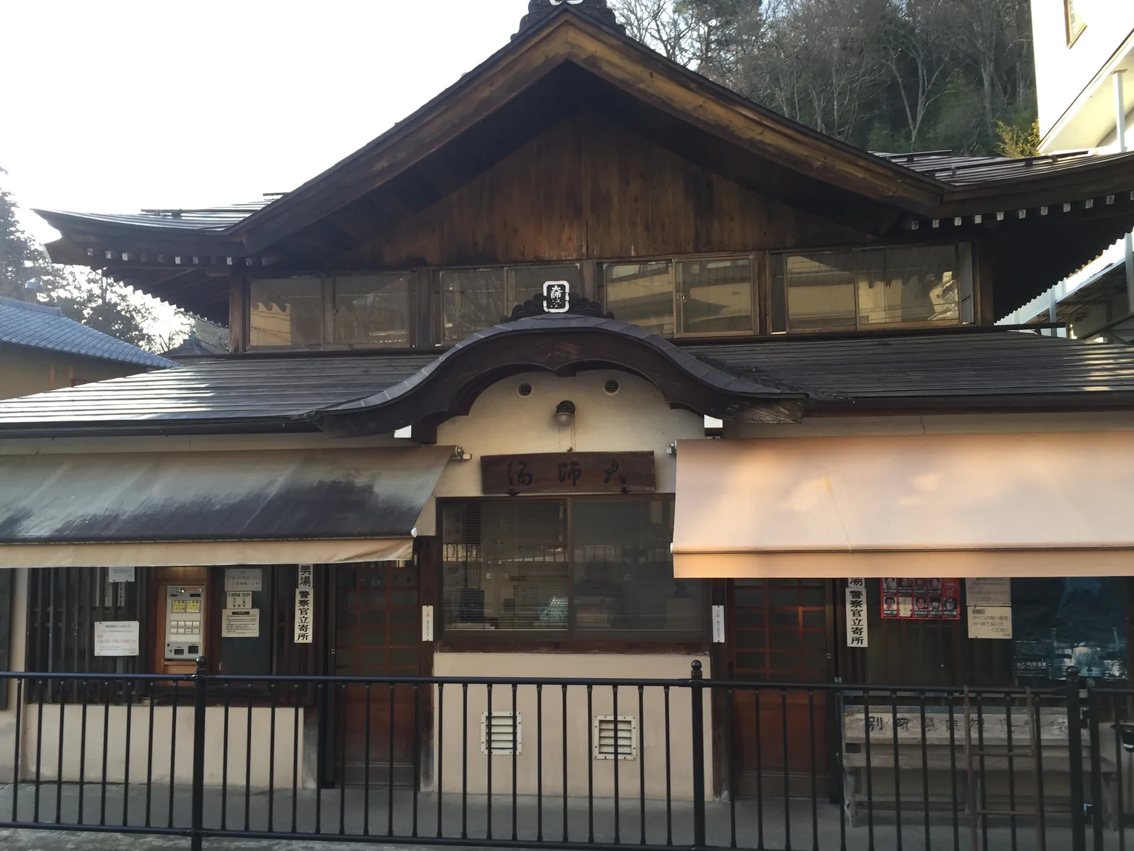 別所溫泉的外湯「大師湯」正面：扁額橫書右起「大師湯」，軒唐破風的玄關上再疊一層切妻的上屋，二樓是一排玻璃窗，左右兩塊縱向木牌分別寫「男湯　警察官立寄所」與「女湯　警察官立寄所」，左端是券售機