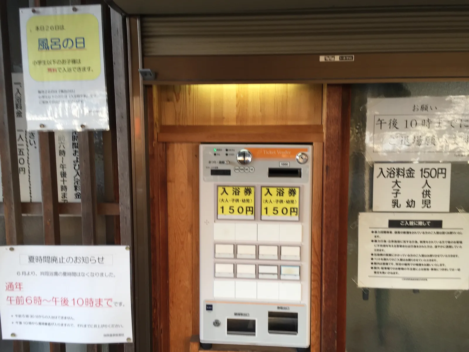 大師湯門口的券售機與告示：黃色按鍵寫著「入浴券（大人・子供・幼児）150円」，右側白紙重複同一個價，左側告示寫「夏時間廃止のお知らせ／通年　午前6時〜午後10時まで」並落款別所溫泉財產區，上方另一張寫當天二十六日是「風呂の日」小學生以下免費