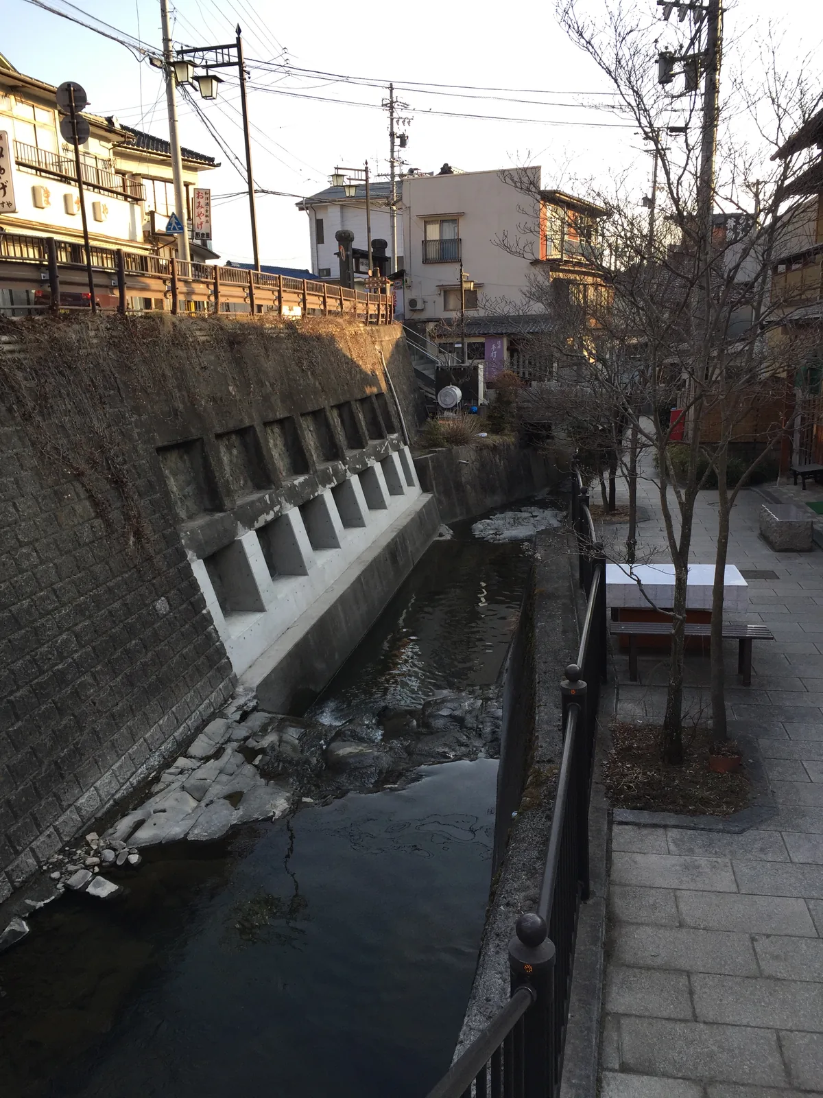 別所溫泉街中央的湯川：兩岸的旅館貼著崖頂蓋，左岸是石砌加混凝土的擋土牆、白色扶壁一根一根排開，右岸是石板步道與長椅，三月水量很小，河床的岩石整片露著