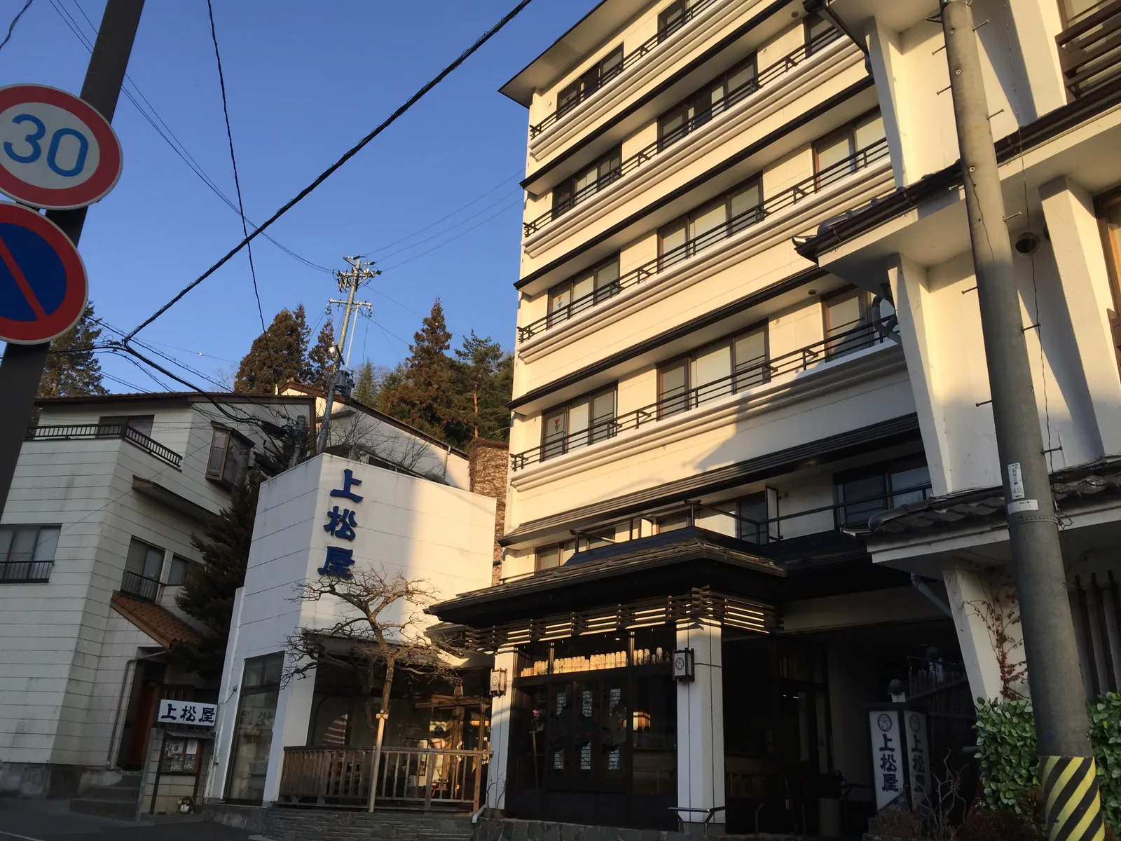 溫泉街裡的旅館「上松屋」：六層的白色建物貼著山腹立起來，正面玄關是唐破風的木造車寄，簷下掛著一排行燈，左側白牆上縱書著店名，背後是被朝陽照到的針葉樹山坡