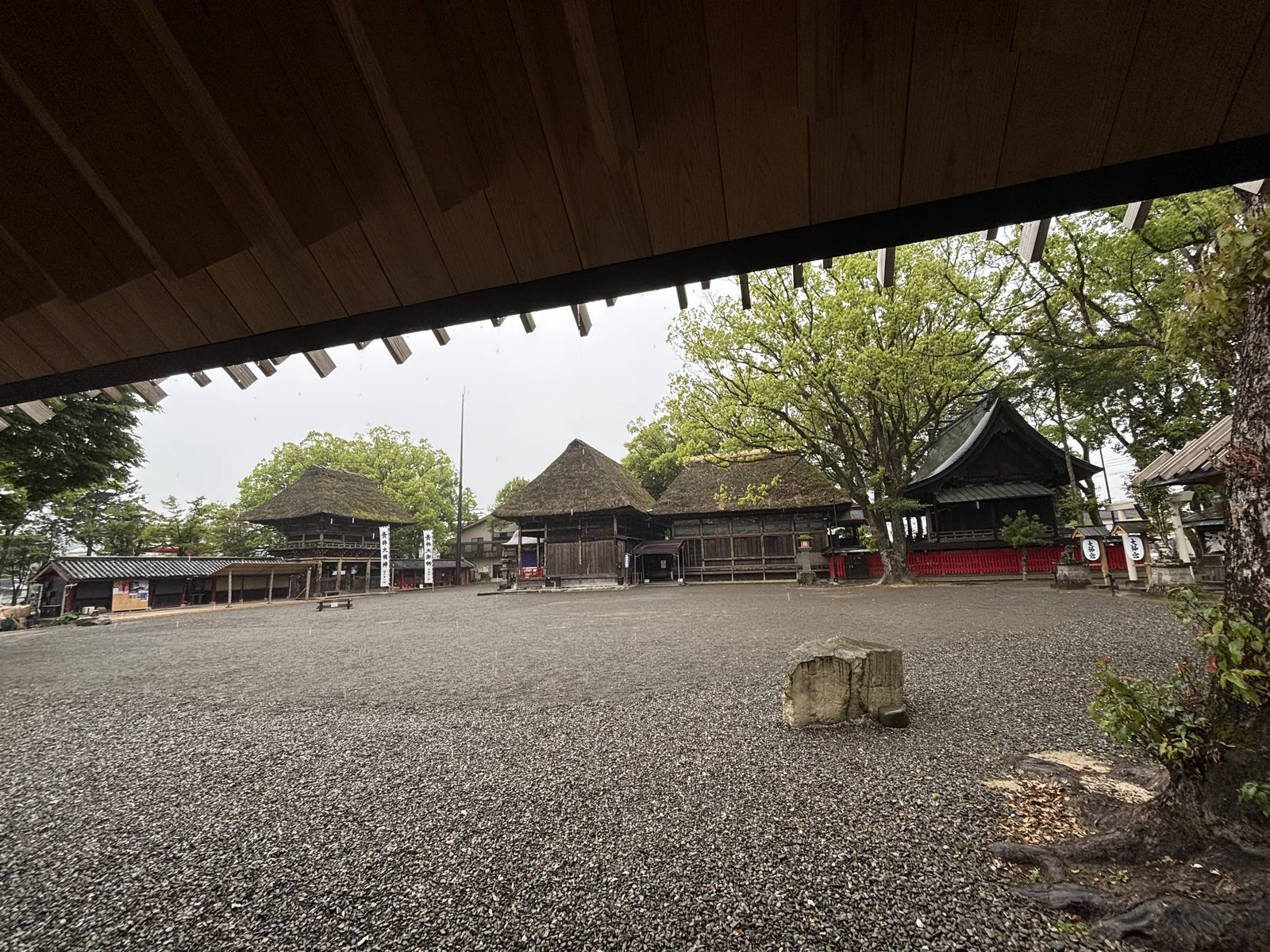 從紀念館軒下望向社殿群，雨中的碎石前庭與一排茅葺屋頂