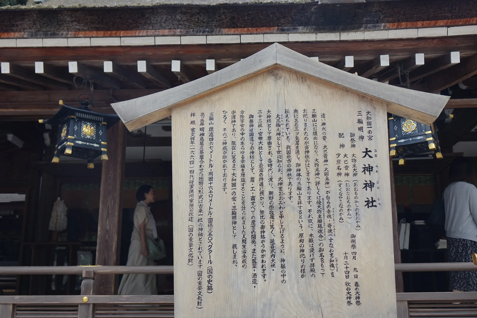 大神神社拜殿前的木製由緒板，直排刻著御祭神、御例祭與由緒全文，背後是拜殿的木構與吊燈籠