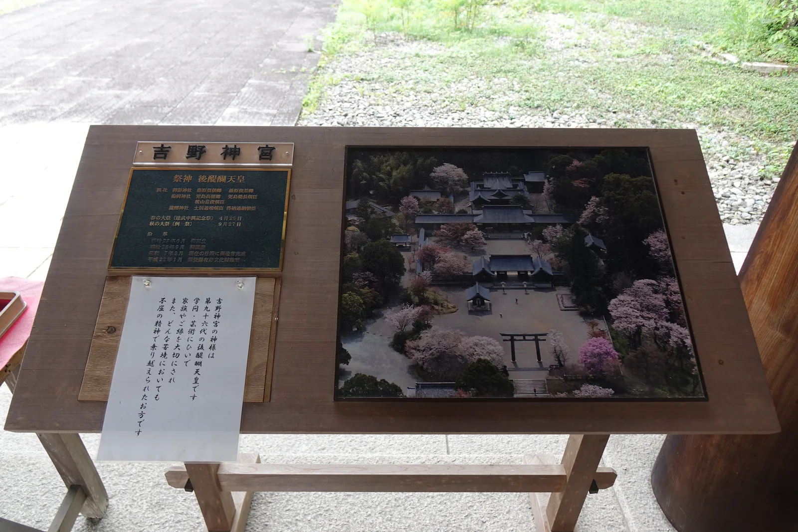 吉野神宮的屋外案內台，木台上並排著社號板、綠底金字的沿革銅板與境內空拍照，左下角壓著一張手寫掲示