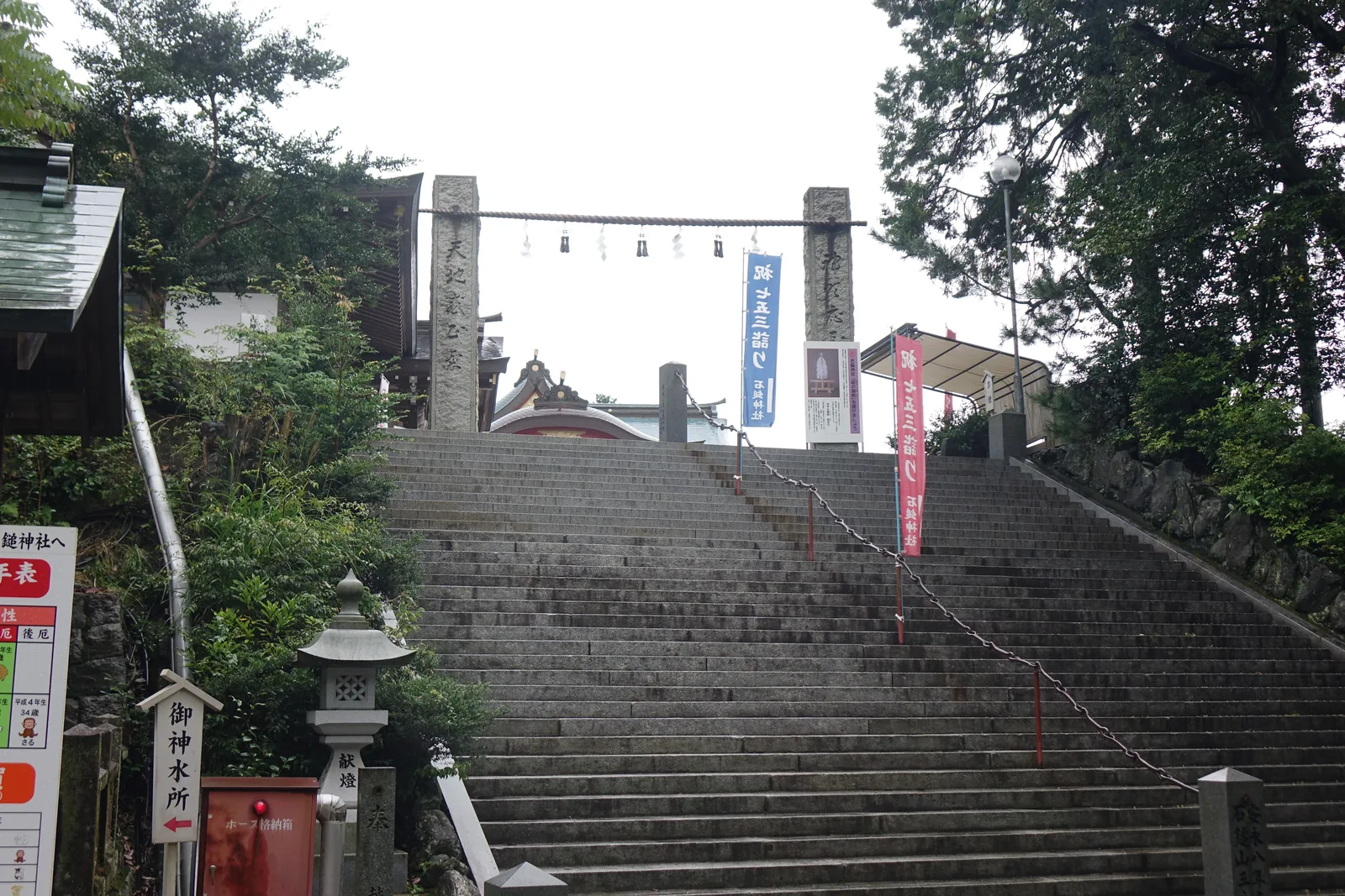 石鎚神社的石造注連柱，兩根方柱之間橫著注連繩垂六束紙垂，石段中央那條鐵鏈一路往上，兩側插著紅藍兩色的七五三詣り幟