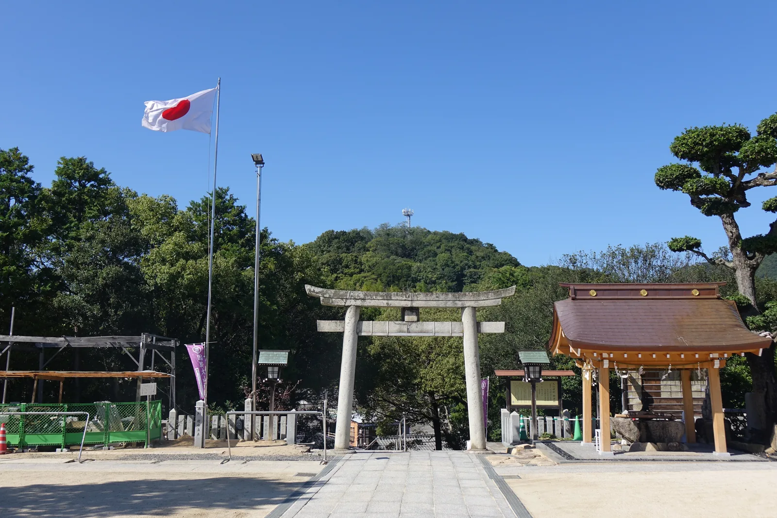 大山神社境內入口的石造明神鳥居與日章旗，右手是木造的手水舍，鳥居正上方背後山頭上的電視塔就是天狗山，左側是造替工事的綠色圍籬