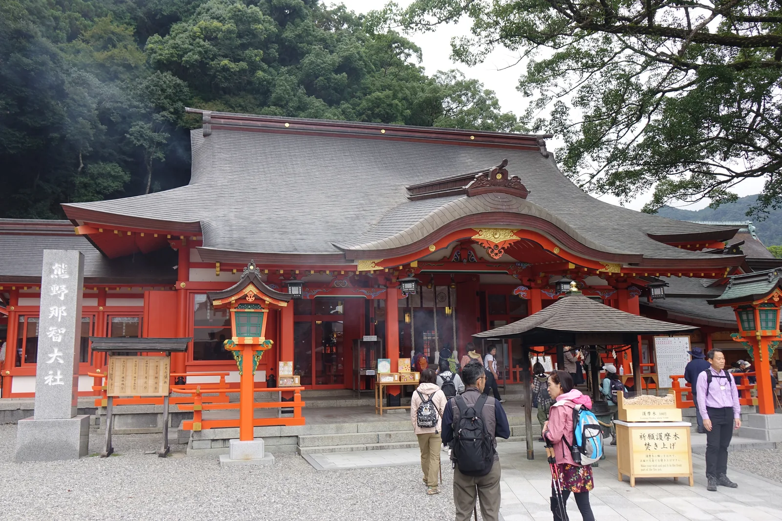 熊野那智大社的禮殿正面，入母屋造、檜皮葺，正面一個大唐破風向拜，朱漆的柱配白壁與金色彫飾，前方小亭裡燻著護摩的煙