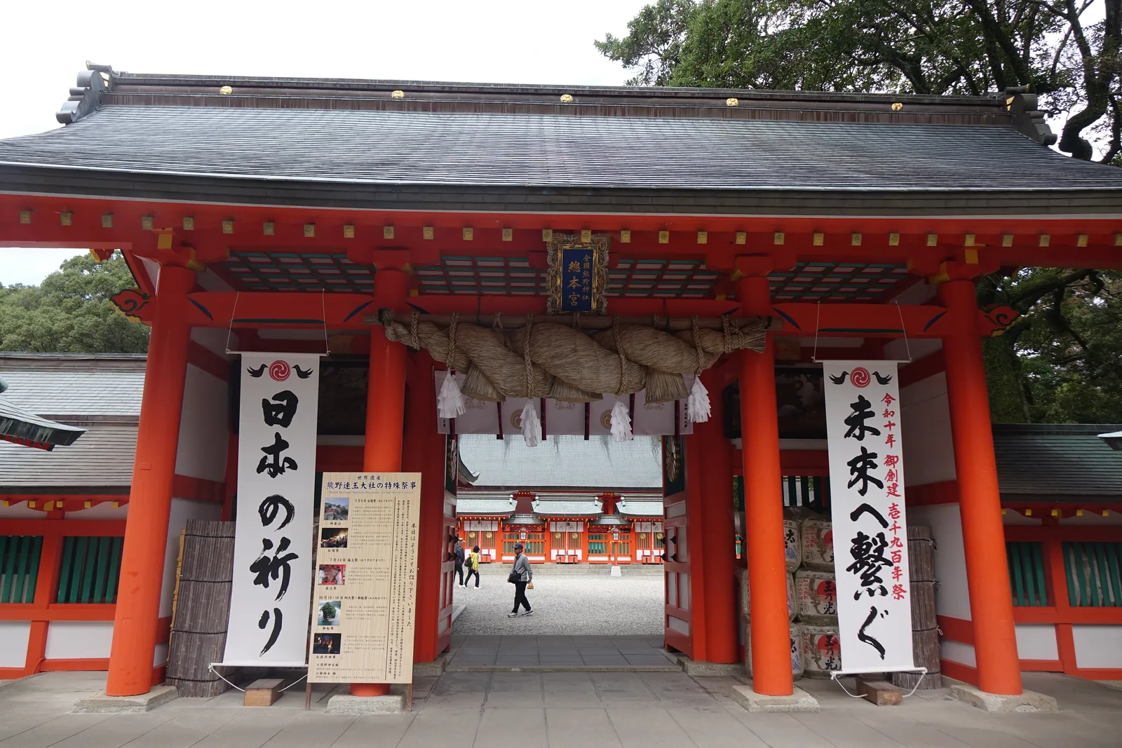 熊野速玉大社的神門（神社的正門），朱漆的柱、黑瓦的屋頂，門楣下橫著一條注連繩，兩側垂下寫著「日本の祈り」（日本的祈禱）「未來へ繋ぐ」（連向未來）的白幟