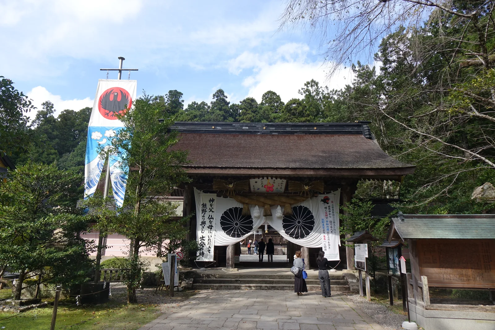 熊野本宮大社的神門，素木與檜皮葺的厚屋頂，門前橫著一條粗注連繩，底下垂兩片大白布，各印一個放射狀的黑色菊紋