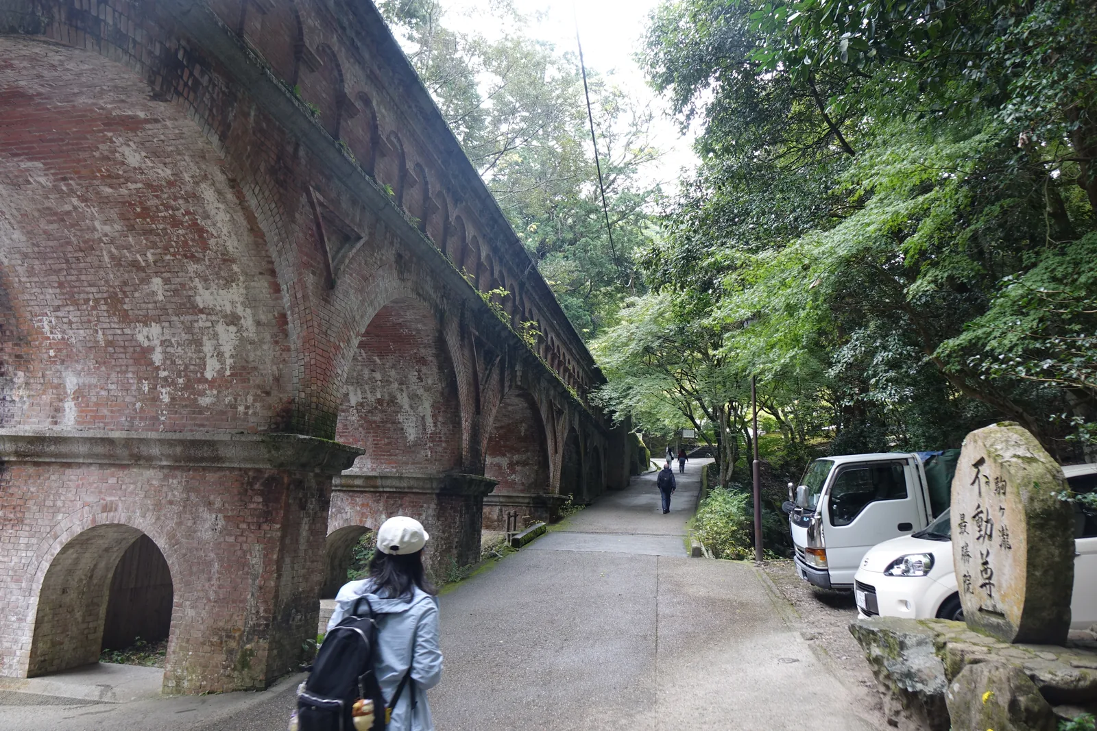 南禪寺水路閣的側面：紅磚砌的連續拱廊斜斜退入樹叢，橋上有水在流，右側立著刻有「駒ヶ瀧　不動尊　最勝院」的石碑