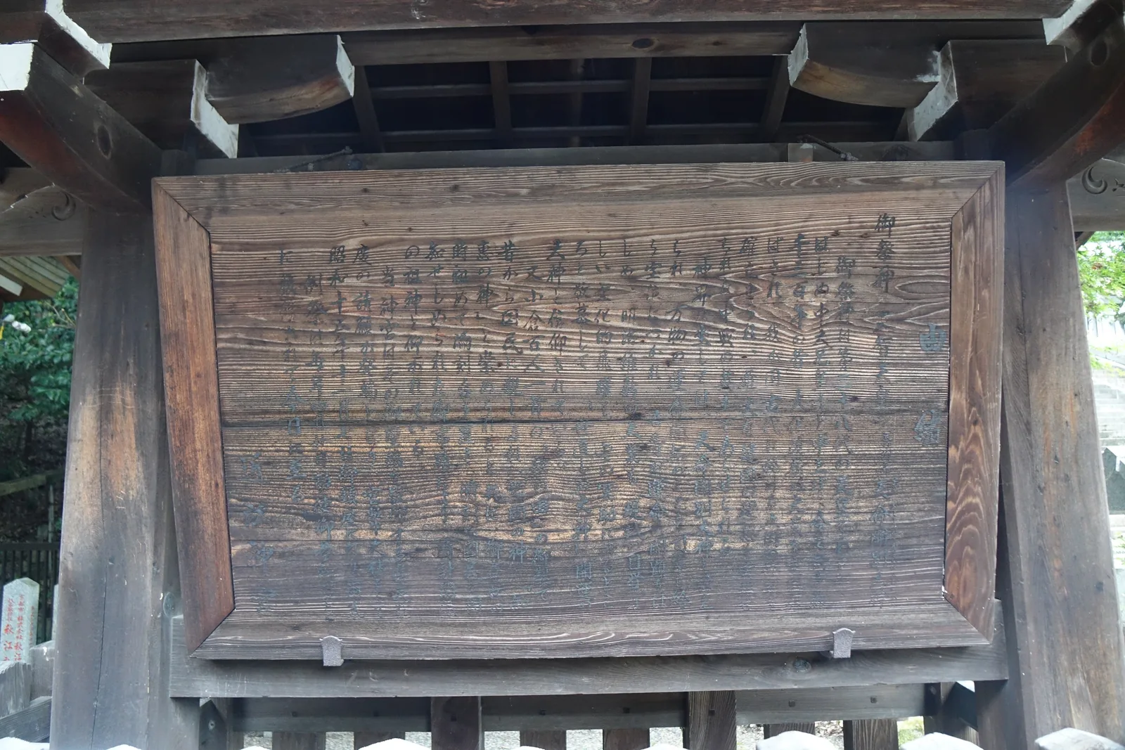 近江神宮境內掛在小屋頂下的木牌，標題刻著「御祭神」與「由緒」，內文的刻痕已被風化磨平，木紋浮起壓過筆畫