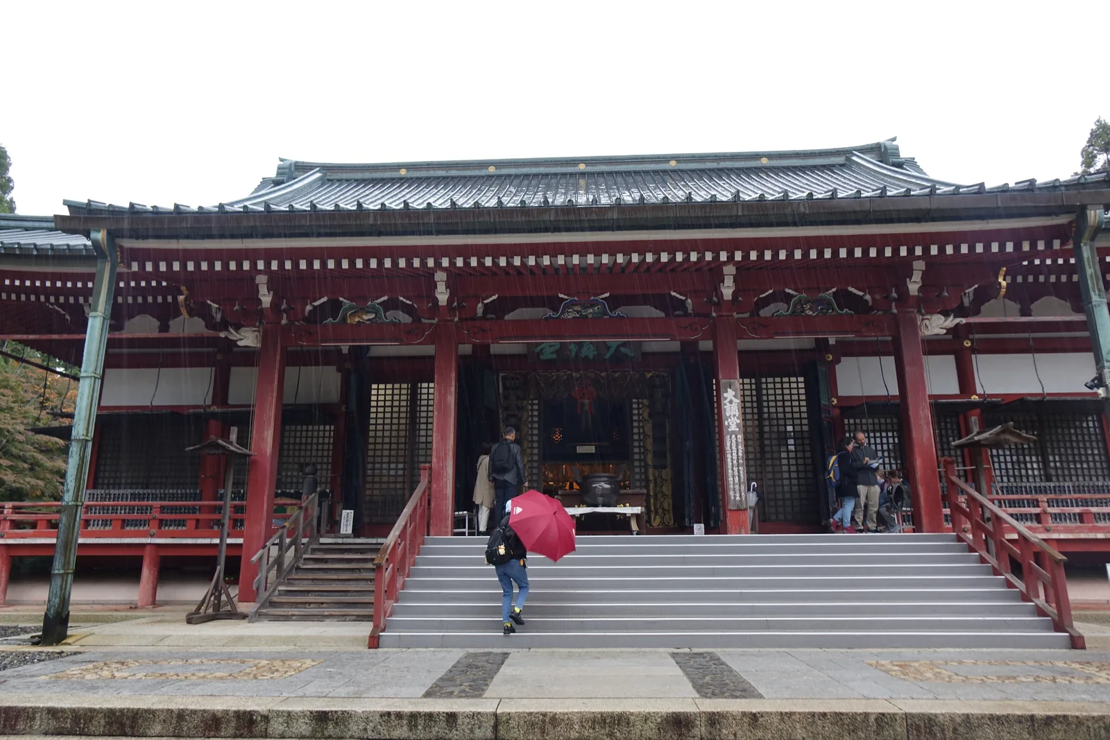 比叡山延曆寺東塔的大講堂正面：入母屋造的大堂，朱漆的柱與白牆，正面一排綠色格子門，前方是石階，雨中