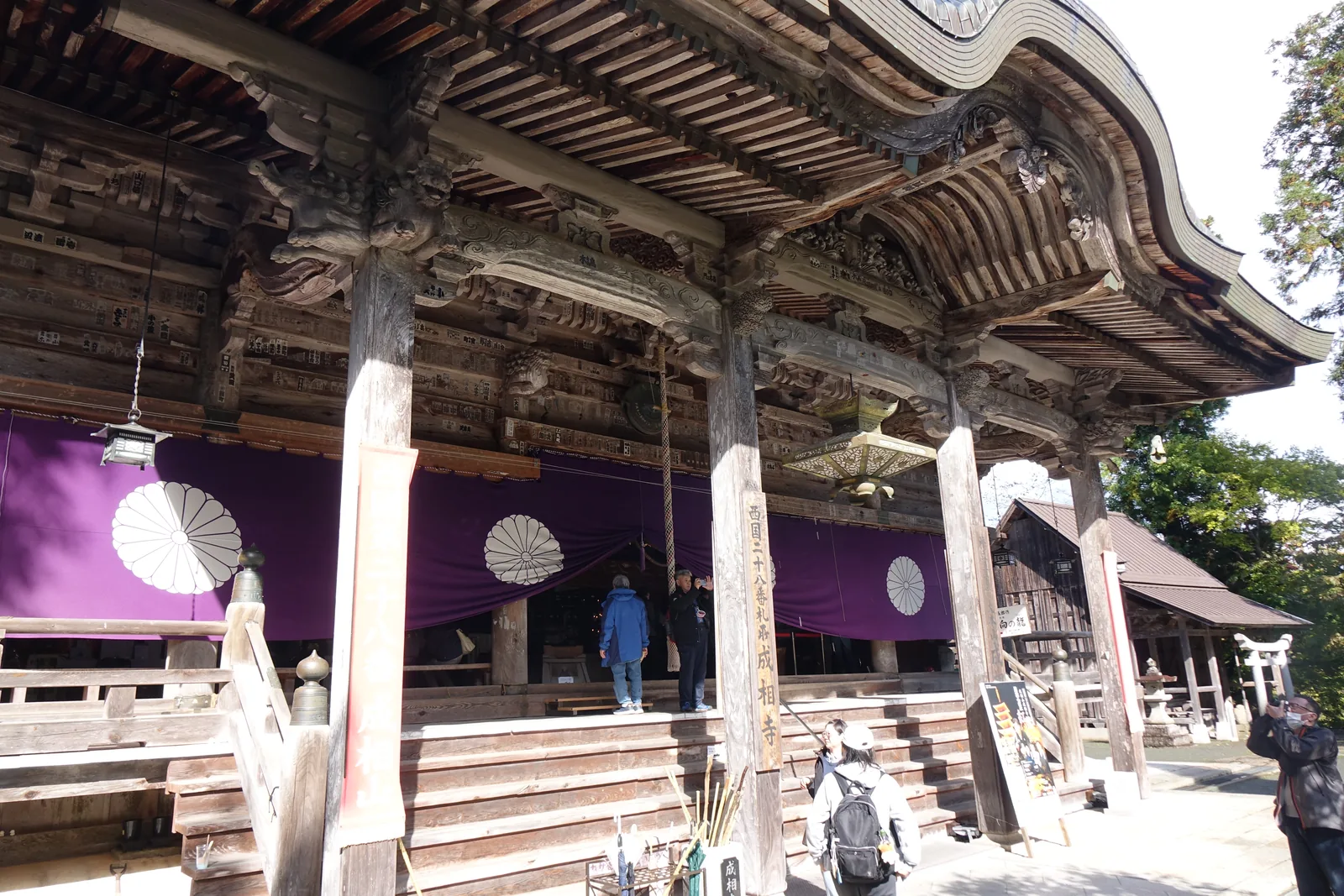 成相寺本堂正面：深色木造的入母屋，屋簷向前挑出一座唐破風，柱上墨書「西國二十八番札所　成相寺」，前方是石階與參拜的人