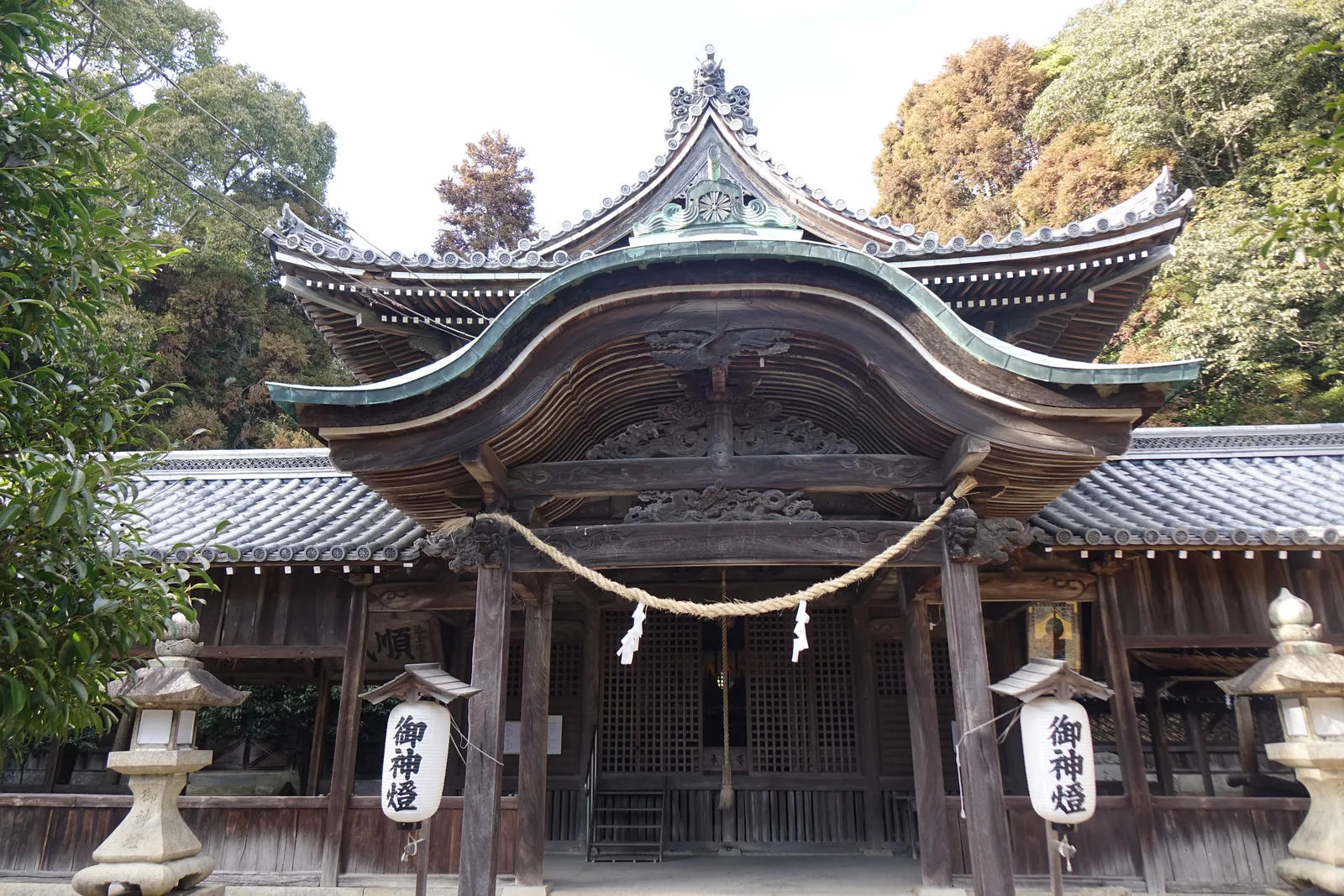 大避神社拜殿正面：深色木造，屋簷向前挑出一座很大的唐破風，注連繩橫過整個開間，兩邊掛著寫「御神燈」的提燈