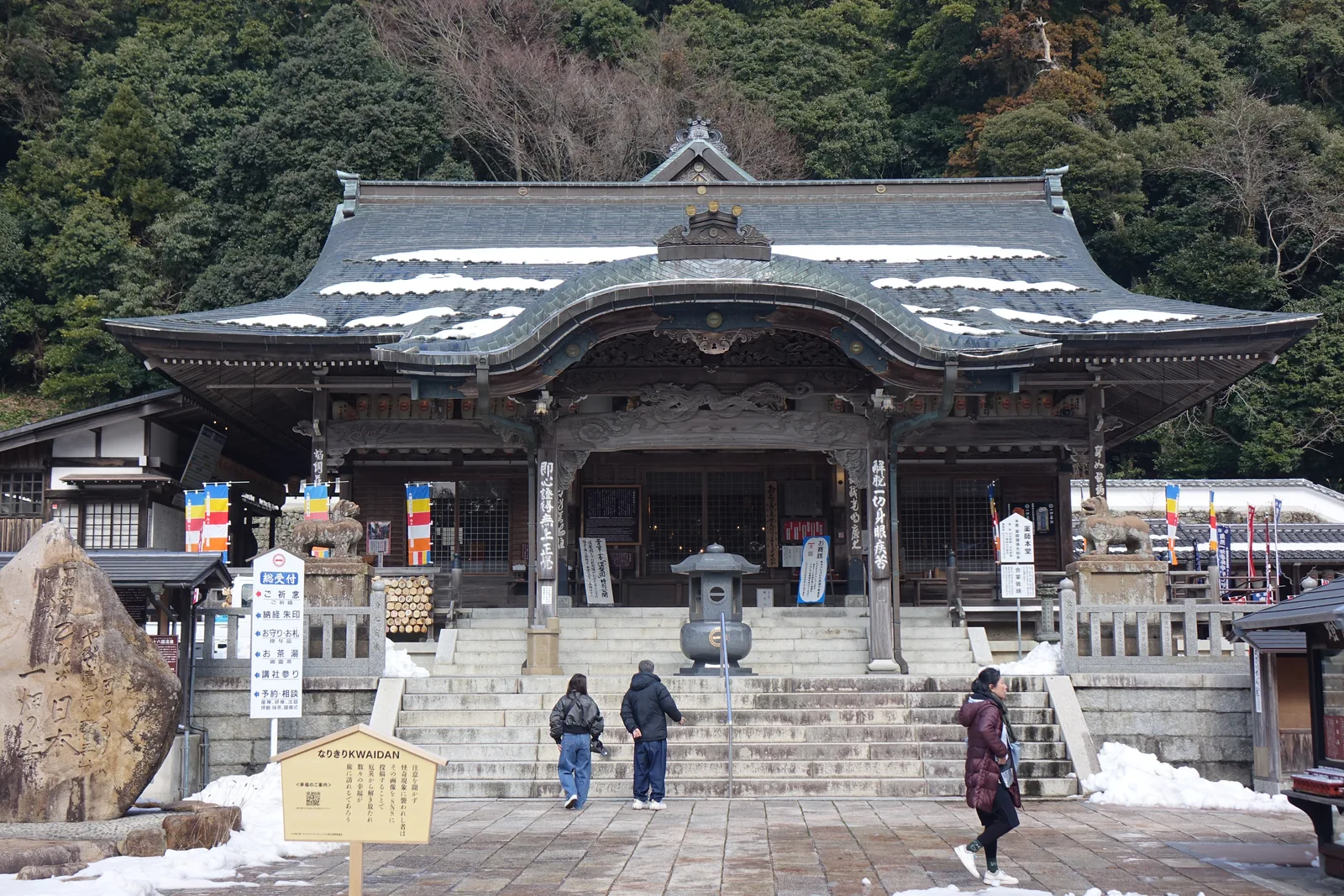 藥師本堂正面。大屋頂上留著殘雪，正面的破風層層挑出，簷下的虹梁盤著龍。右側的柱牌刻「解脫一切身眼疾苦」。左前方自然石上刻的是這座寺的巡禮和歌。石階前那具青銅香爐後面，就是總受付。