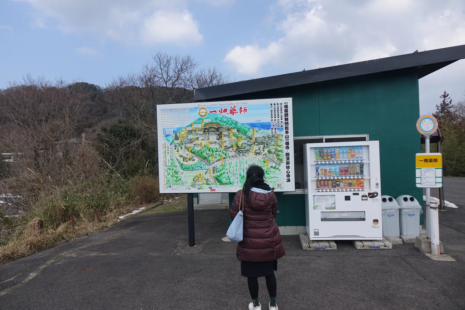 山上停車場的一角，兩塊看板與一台自動販賣機，一個人正在讀圖。這是全部照片裡唯一能看見山上聚落尺度的一張：路是柏油的，房子是鐵皮的，二月的路邊還有積雪。
