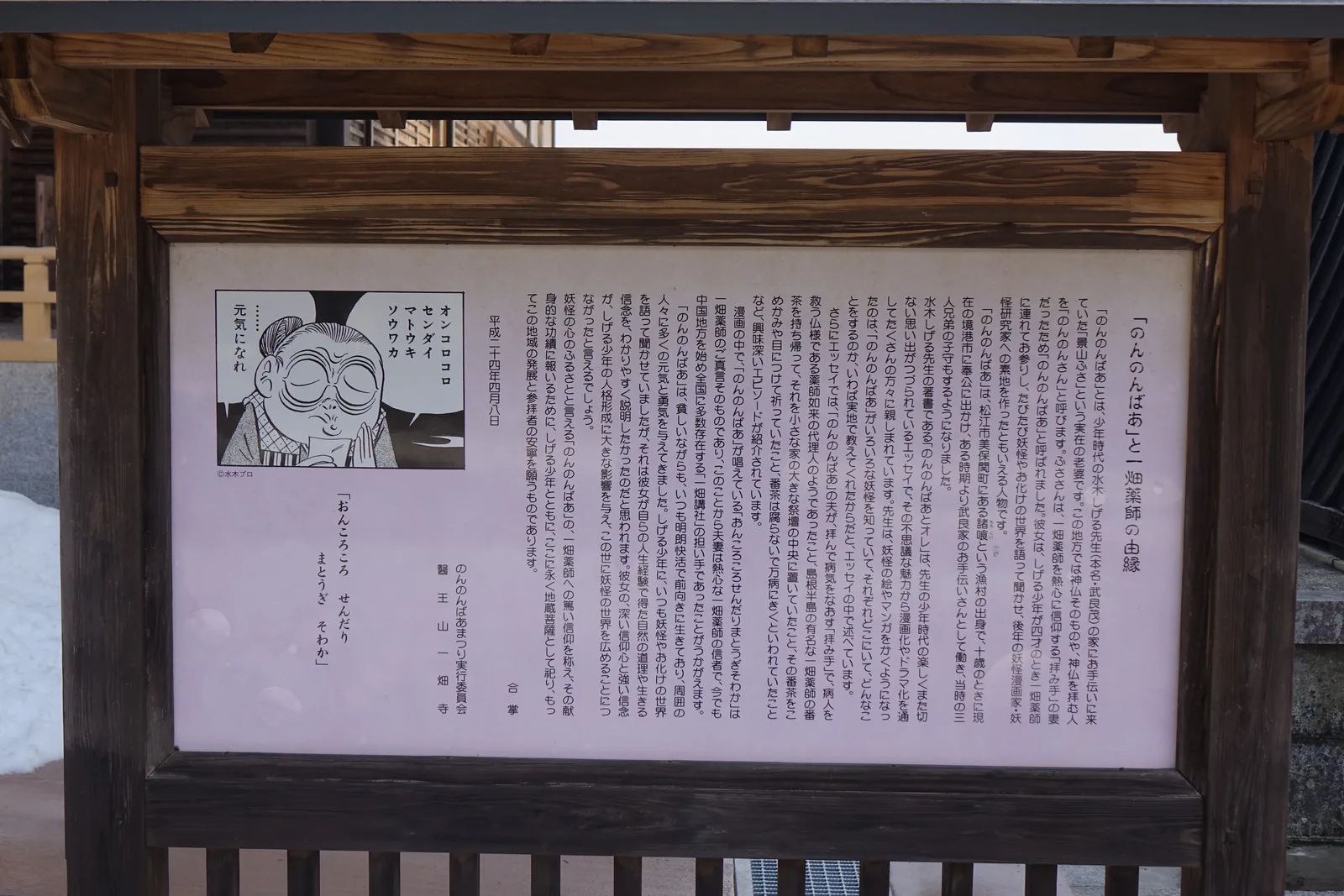 木框裡的說明板，落款平成二十四年四月八日，署名「のんのんばあまつり実行委員会」（のんのんばあ祭典執行委員會）與醫王山一畑寺。板上引了水木しげる散文裡的細節：她丈夫像藥師如來的代理人、從一畑藥師帶回的番茶放在祭壇正中央、番茶抹在太陽穴與眼睛上、據說不會腐壞。左上一格漫畫裡，のんのんばあ正念著藥師的真言。