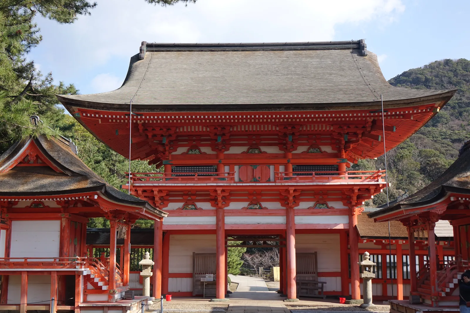 樓門正面。二層、塗朱，屋頂是檜皮葺（hiwada-buki，用檜樹皮層層鋪成的屋頂），下層三開間而只有中央一間通行；上層有帶欄杆的緣，梁上一排彩色的雕刻。門後可以看見日沉宮的檜皮屋頂與更遠的松林。門的左右各伸出一道白牆廻廊。