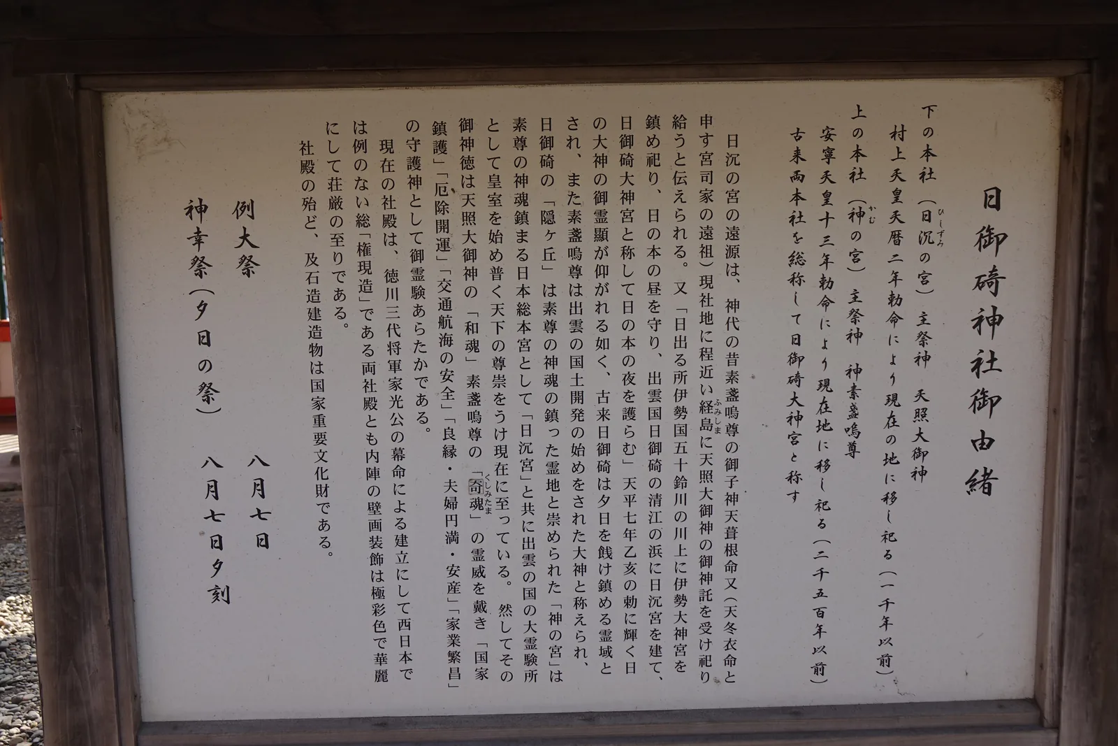 木框裡的直書立牌，即由緒板（yuisho-ban，記載神社來歷的解說牌）。開頭兩行把上下兩座本社的祭神與遷座勅命並列，第三行寫「古来両本社を総称して日御碕大神宮と称す」，自古兩座本社合稱日御碕大神宮。中段抄了天平七年乙亥之勅——伊勢守晝、日御碕守夜。末段記現社殿是德川家光幕命所建的總權現造，例大祭八月七日、神幸祭（夕日の祭）同日傍晚。