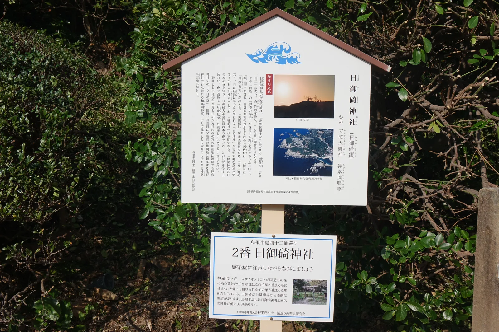 屋形的白板，島根半島四十二浦巡り再發現研究會製作。上半抄《出雲國風土記》寫御前濱「広さ一百二十歩あり。百姓の家あり」——寬一百二十步，有百姓的房子——並記海人採的鮑魚是名品；下半列出三場祭禮。柱下另掛一塊小板：「2番 日御碕神社」，寫神蹟隠ヶ丘的柏葉傳承，並註明參道從燈塔停車場往南進入。
