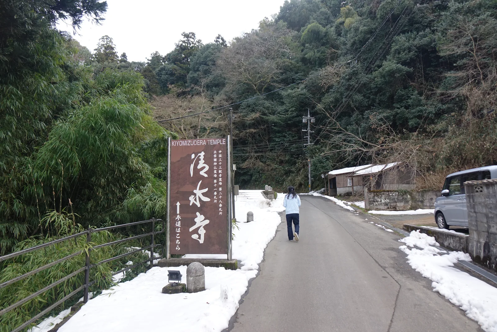 縣道從谷口往上，路面濕、兩側堆著鏟起的雪。左側棕底大看板：上緣「KIYOMIZUDERA TEMPLE」，中央大字「清水寺」，右側直書「中国三十三観音霊場 第二十八番札所／出雲國神仏霊場 第十一番札所」，下方箭頭與「參道はこちら」。看板腳邊一具小石佛。