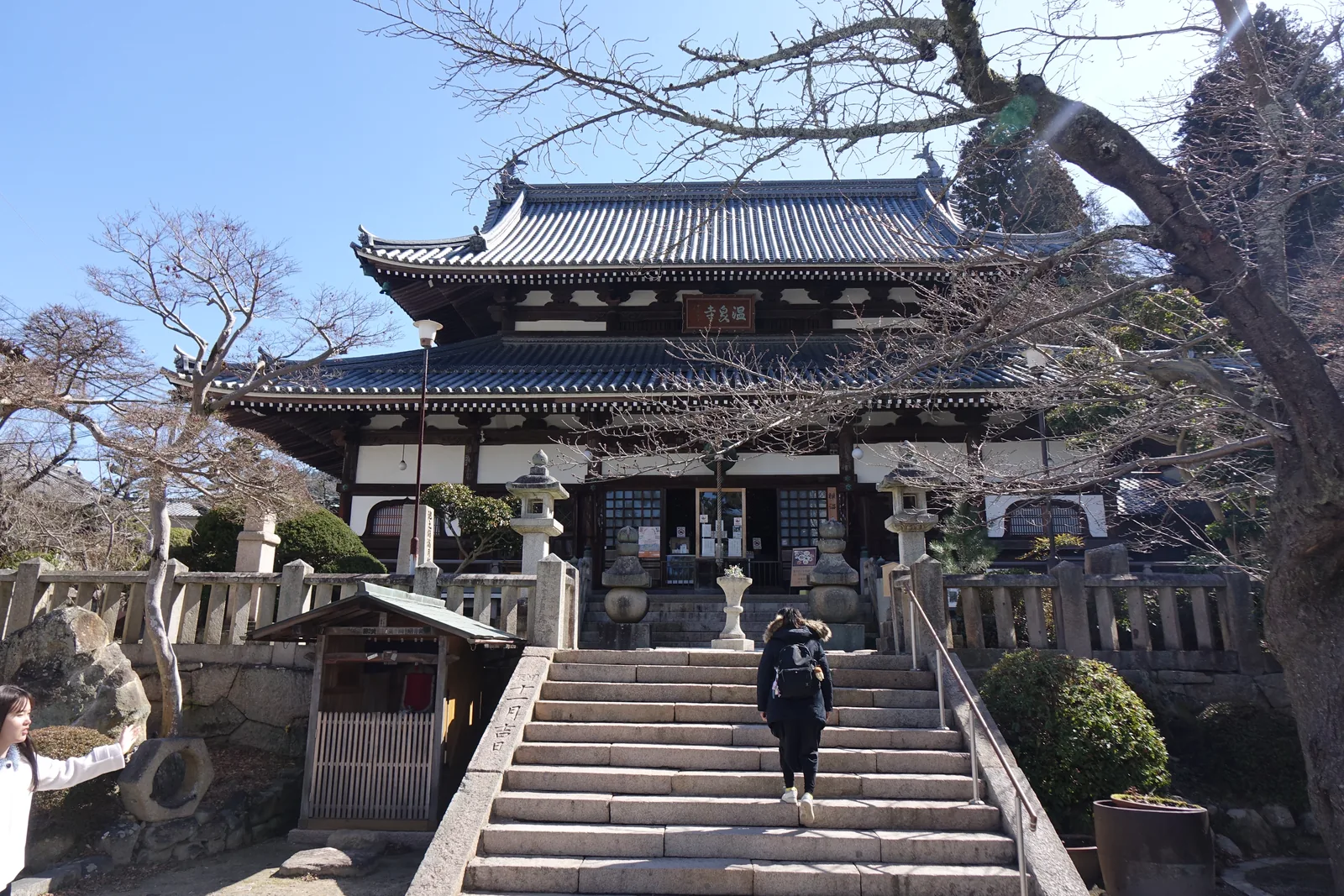 本堂（藥師堂）正面，隔著一段石階看上去。二重的本瓦葺（平瓦與丸瓦交互鋪成的屋頂）入母屋屋頂，正面伸出向拜（堂前突出的一段簷下空間），上層簷下懸著紅底金字的扁額「温泉寺」。石階兩側是擬寶珠的石欄干，右側欄干的側面刻「甲辰　十二月吉日」；階前左右各一座石燈籠。左下角一座覆屋，裡面是紅衣的石像。現在的這棟建築是天明二年（1782）建的，比它下面的石階年輕，比它上面的扁額年輕。