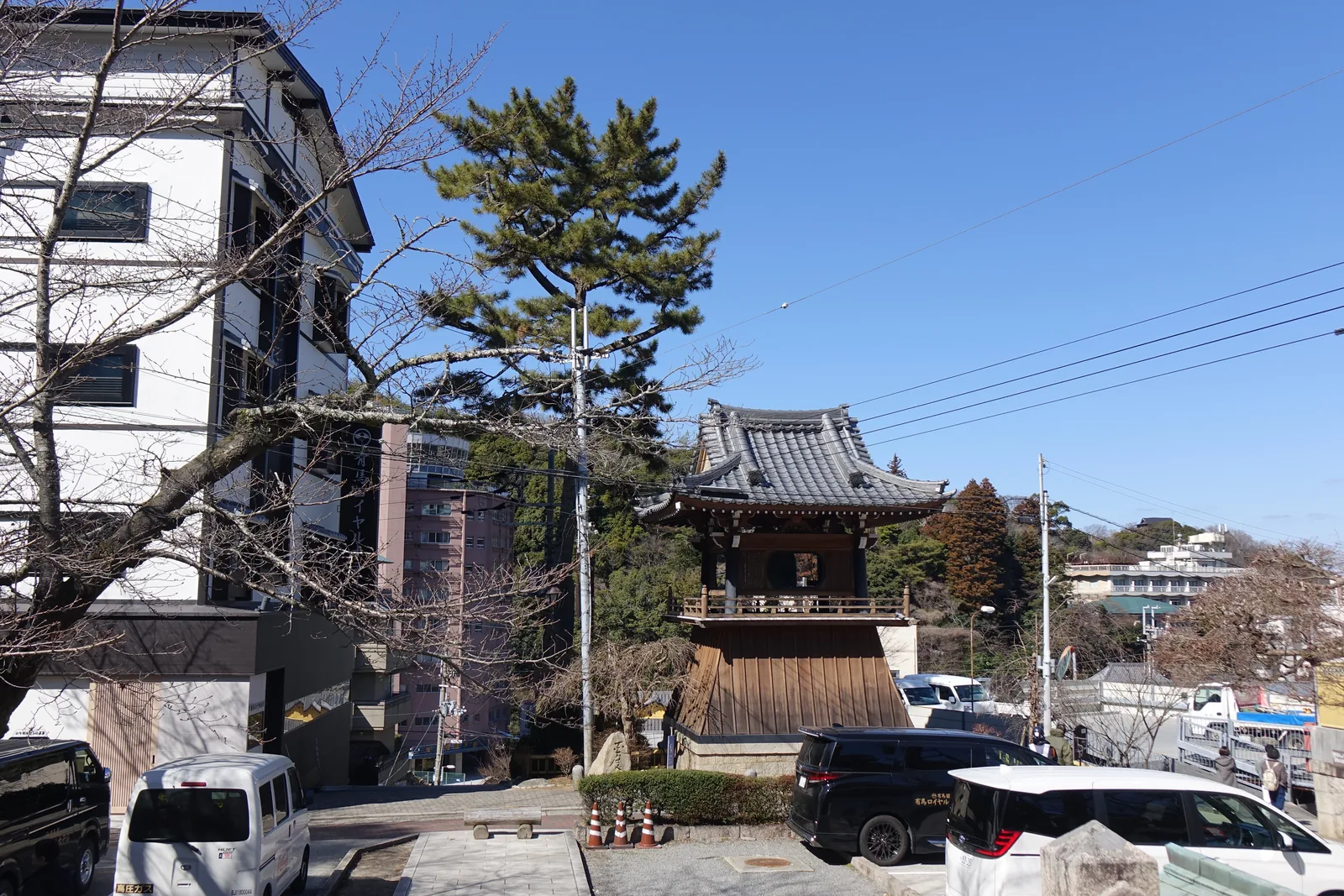 從停車場看鐘樓。本瓦葺的入母屋屋頂，下部是向外斜張的袴腰板壁，立在石砌的平台上。左邊是四五層樓的旅館，右邊是往山上去的樹林與更多旅館，前景是電線、三角錐與停著的箱型車。除夕夜這口鐘會響過整條溫泉街，寺方說觀光客也可以撞。