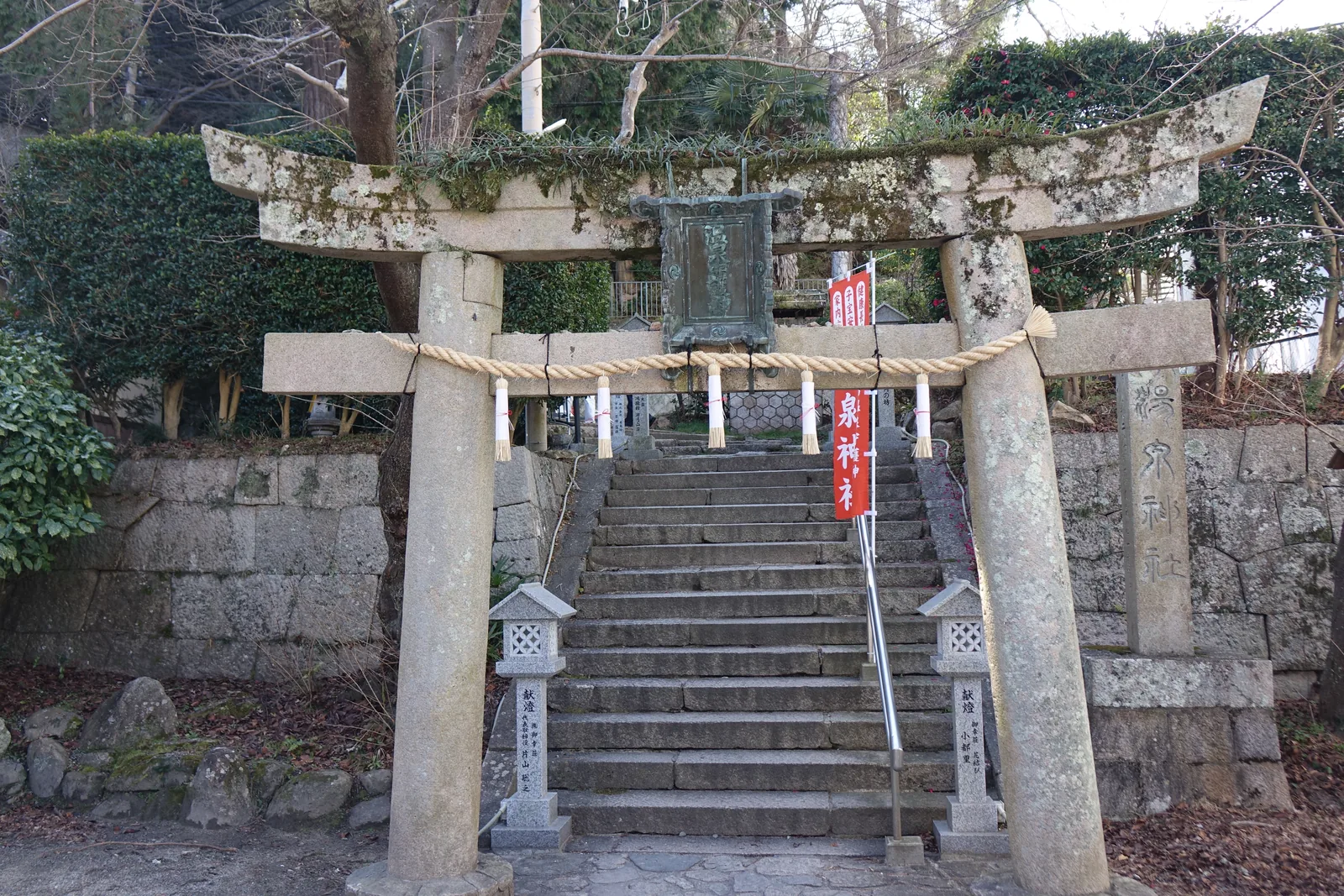 石造明神鳥居，笠木與島木上長著苔與草，中央懸一面綠鏽的青銅扁額，右側另立石標刻「湯泉神社」。注連繩垂五條紙垂，右柱旁掛一面紅幟。兩柱腳各一座石燈籠，竿刻「獻燈」與奉納者名。背後石階筆直上去，兩側切石積的擋土牆。拍攝位置在溫泉寺那一側的平台上——這道鳥居與那間寺之間只有一百一十公尺。