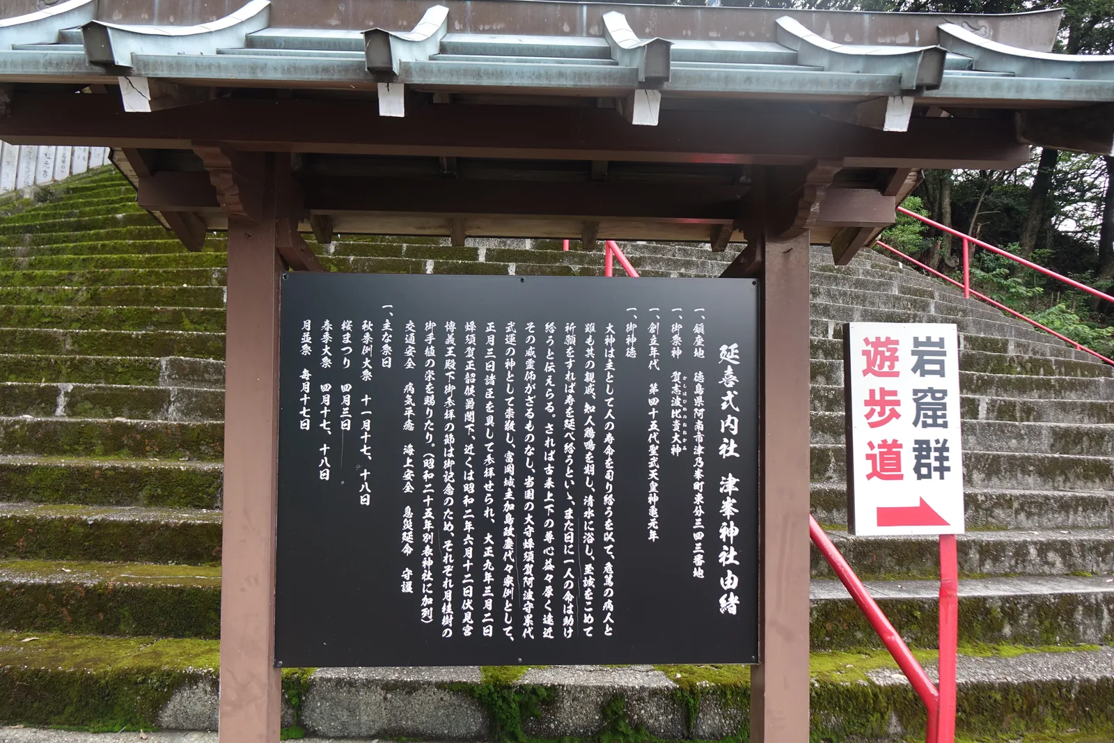 津峯神社石段下的黑底由緒板，立在木造小屋簷底下，抬頭寫「延喜式内社　津峯神社由緒」，右側是紅底白字的「岩窟群　遊歩道」指標