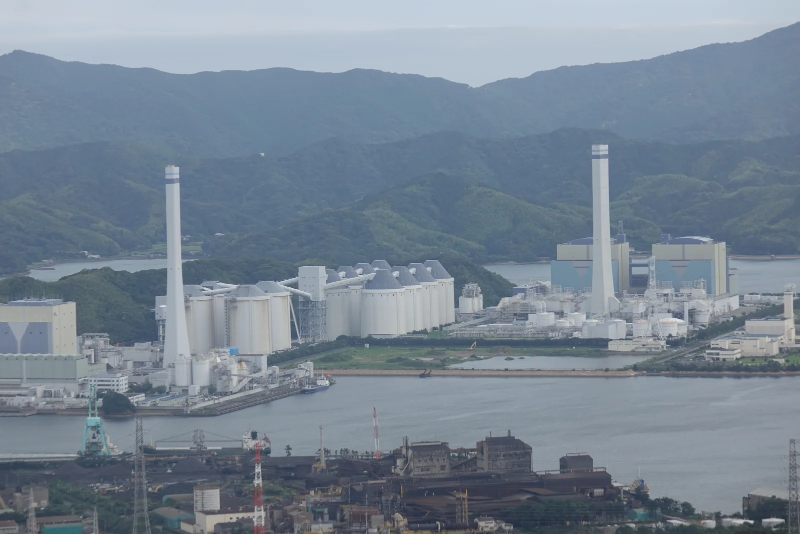 從津峯山展望台往東南看：橘灣岸邊立著幾支巨大的煙囪與一整片廠房，那是二〇〇〇年運轉的橘灣火力發電所