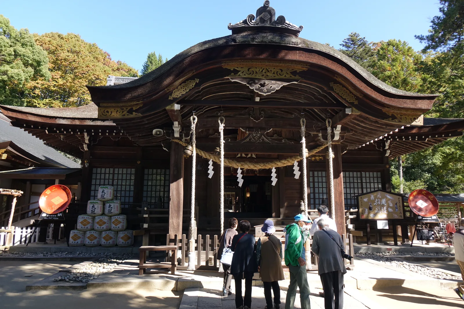 拜殿正面。入母屋造（上半歇山、下半四坡的屋頂），正面伸出一段深深的向拜（殿前突出的一段簷下空間），簷口做成軒唐破風（簷前緣做成波形弧線的山牆），素木（不塗漆的原木）不上漆。橫過向拜的注連繩（界分神域的稻草繩）垂著四條鈴緒，兩側掛大提燈，左後方堆著奉納的菰樽。階前站著幾位參拜者。這座社殿落成於大正八年（1919）四月十一日，隔天四月十二日——信玄的命日——就舉行了第一次例祭。
