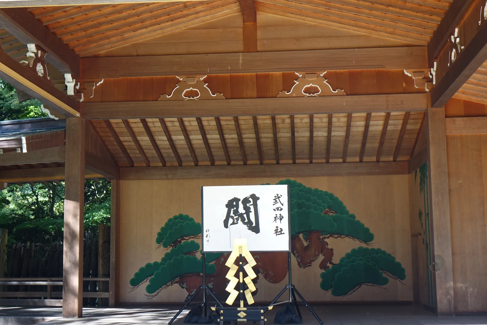 能樂殿內部。鏡板上畫著一株橫展的老松，前面立著一幅寫著「闘」的揮毫，右側添書「武田神社」。台前是一座黑漆的台與金色御幣，台面正中是武田菱的家紋。素木的屋架與欄間的雕花板都還很新——這棟建築竣工於平成十八年（2006）。