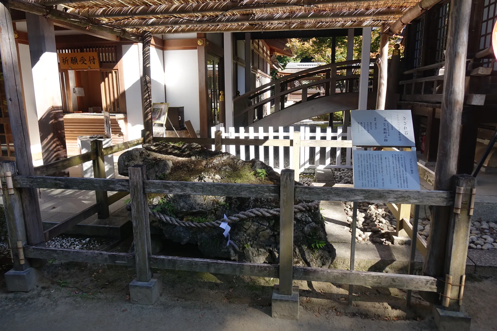 覆棚下的さざれ石，注連繩繞過石身、垂著紙垂（shide，鋸齒狀的白紙垂飾），前後圍著木柵與白木柵。右側兩片解說板寫明產地是岐阜縣揖斐川町、重量將近四噸、由宮司命名為「龜石」。左後方是「祈願受付」的橙色招牌與寺務所，中間跨著一座木造的太鼓橋。