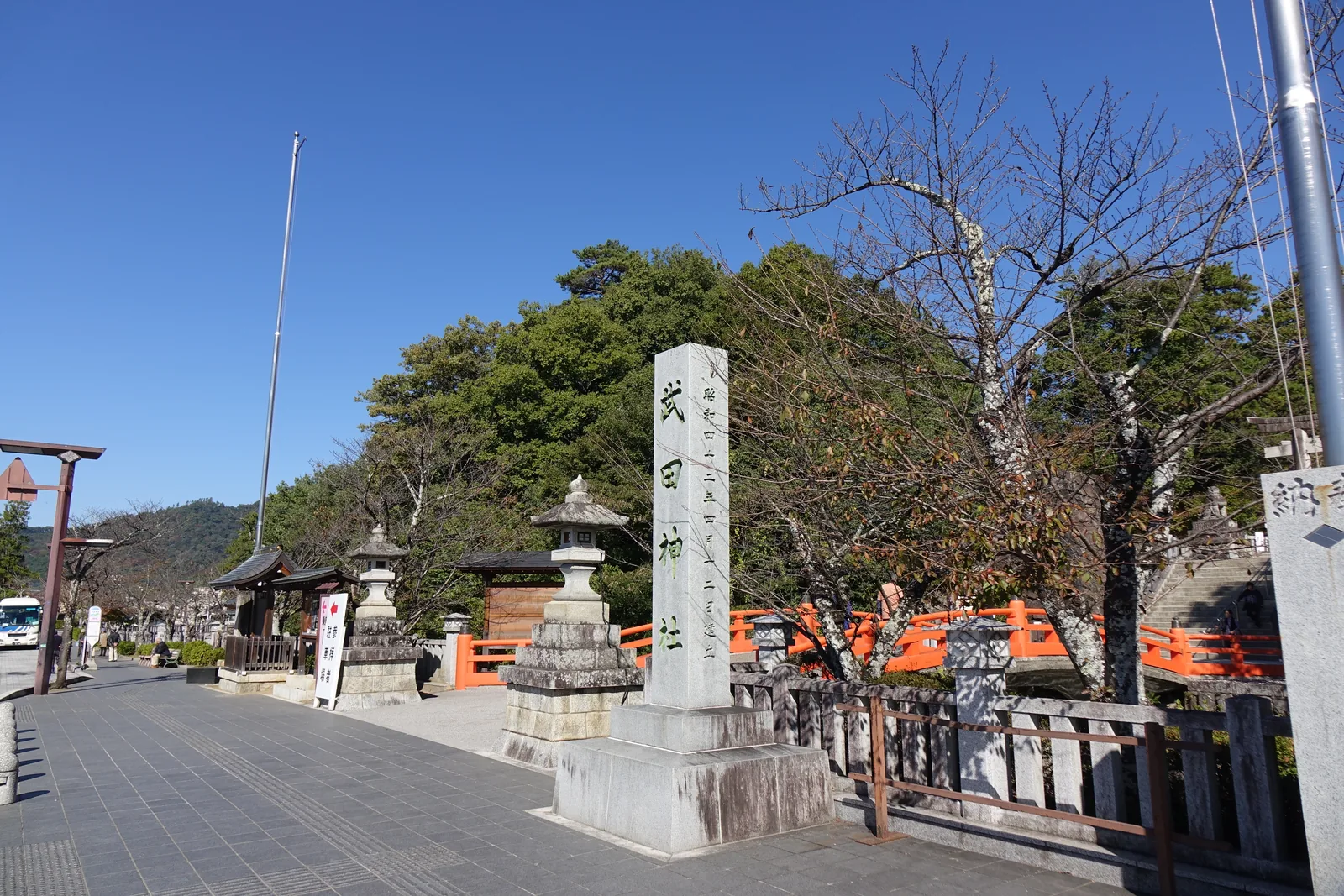 參道口的社號標，正面刻「武田神社」，側面刻「昭和四十二年四月十二日建立」——立標的人挑了例大祭那一天。右側是朱紅的神橋欄干與石燈籠，左側是往驛去的武田通り，遠處是盆地南緣的山。