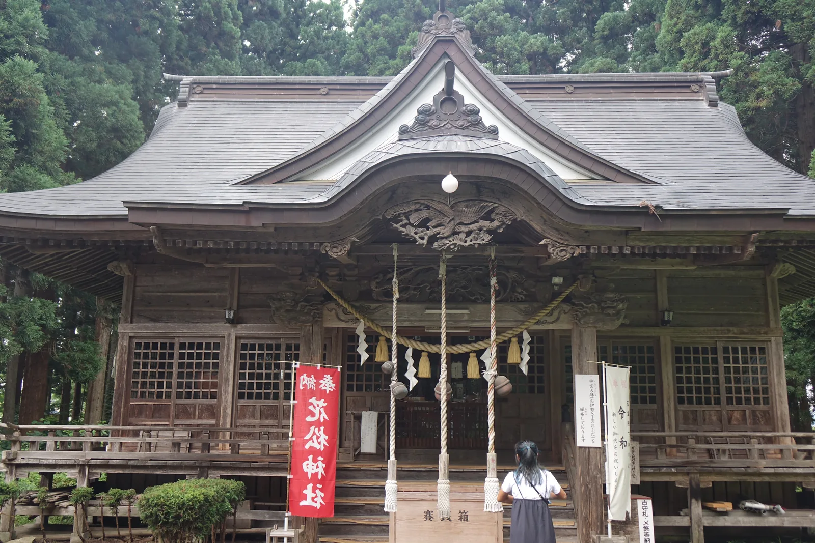 拜殿正面。屋面鋪灰色的金屬板，正脊下方先起一面三角形的破風（はふ，hafu，屋頂山牆邊緣那道斜向的構件），破風之下再挑出一道弧形起伏的簷。弧簷底下懸著一塊雕成鷹的木飾，兩側的橫木上是成對的龍。簷下橫著一條稻草繩，垂下白紙條與兩顆金鈴，兩條白色的繩從鈴上垂到地面。門前左側立著一面紅底白字的幟旗，寫「奉納　花松神社」，右側是白底的幟旗。階前擺著木製的賽錢箱，一個人站在階下。背景是高聳的杉。