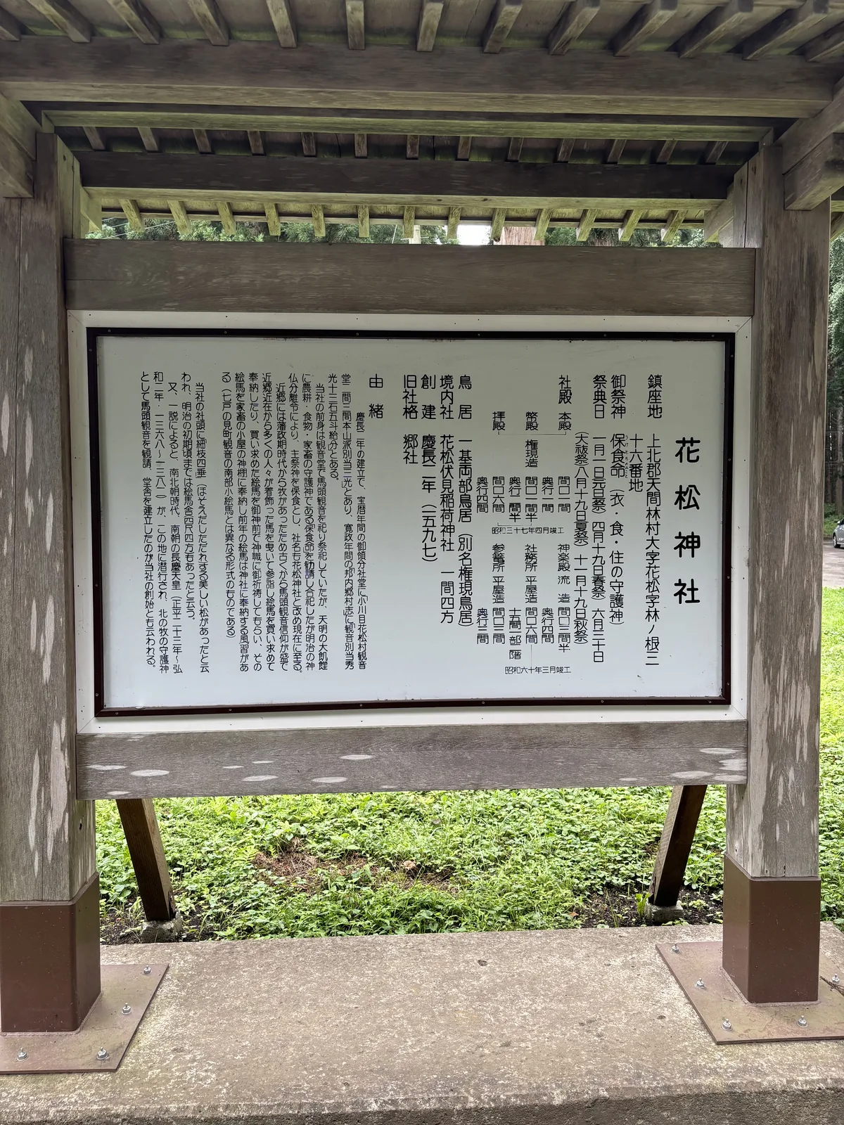 木造的四柱棚架下，一面白底黑字的直書說明板，標題「花松神社」。內容依序列出鎮座地、御祭神、祭典日、社殿的形式與尺寸、鳥居、境內社、創建年、舊社格與由緒。板框是深色的金屬，棚架的木頭已灰化。背後是草地與杉林。