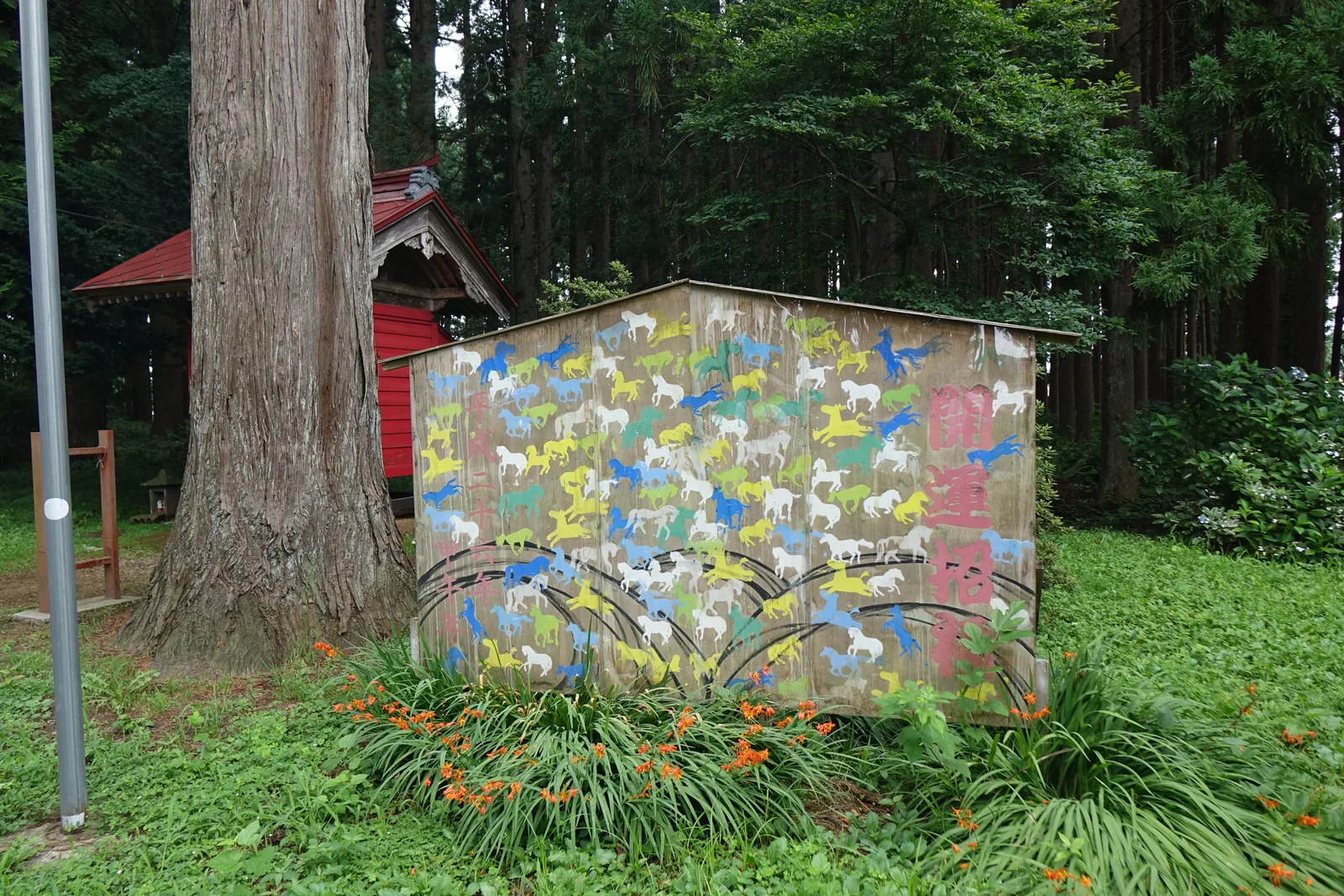 一座金屬板搭成的小屋，外牆上畫滿了幾百匹馬，藍、黃、綠、白四色交錯，全部朝同一個方向。牆的下緣畫著幾道黑色的弧線。右端直書粉紅色的大字「開運招福」，左端另有一行粉紅的小字，寫著年份。小屋左後方是一株樹皮縱裂的巨杉與一座朱漆的小祠。屋前叢生著開橘色花的草。