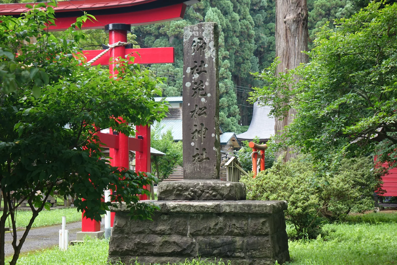 一根方形的石柱立在石砌的基座上，正面直書「鄉社　花松神社」。石柱右側是朱漆鳥居的一角與橫木，左側是枝葉茂密的灌木。更遠處可見境內的朱漆小祠與灰色的屋頂。地面是草。