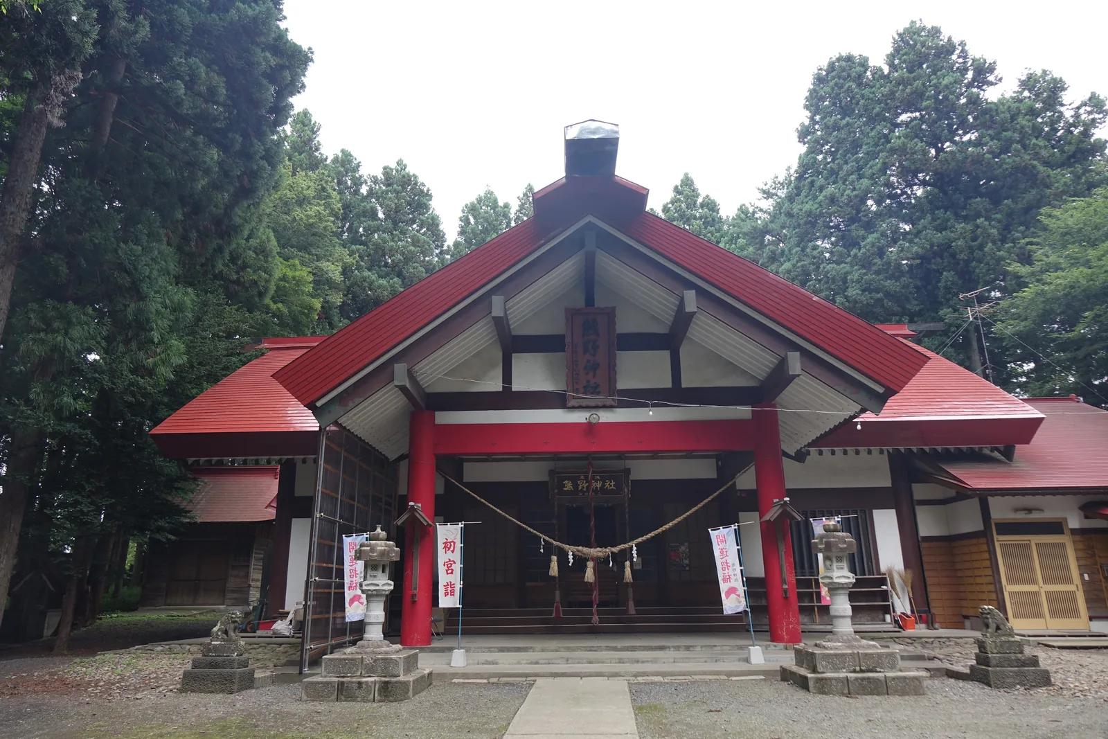 拜殿正面。三角形的山牆正對著參道，屋面鋪紅色的金屬板，兩側各披出一段較低的屋簷。山牆的板面是白色的，中央懸著一塊直書的木匾。屋脊上方立著一支金屬的煙囪。入口的四根柱子漆成紅色，門楣上另掛一塊橫的木匾，底下橫著稻草繩與白紙條。階前左右各立一座白色的石燈籠與一頭石獸，兩側各插一面白色的幟旗。背景是杉。