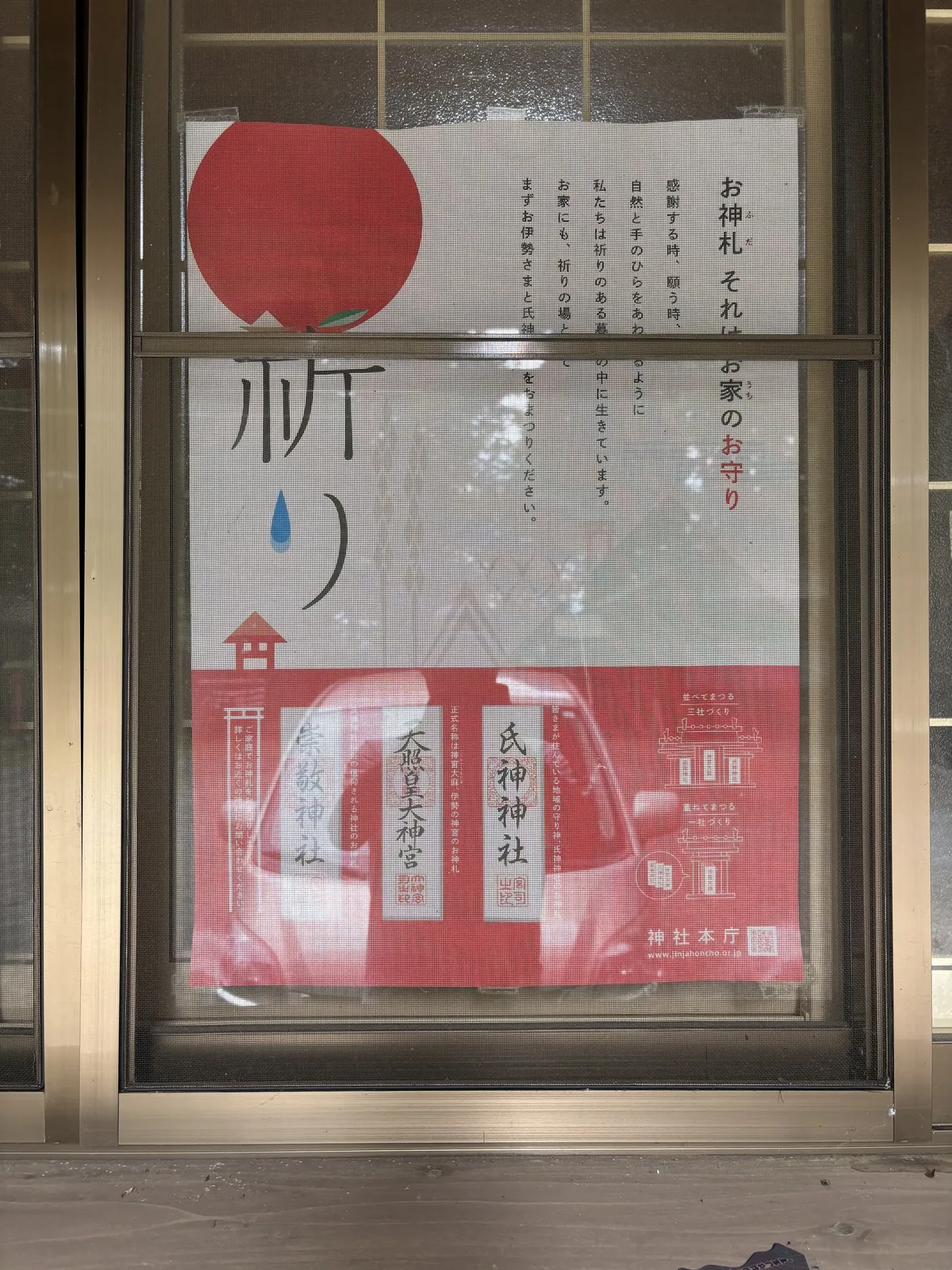 社務所的鋁框玻璃窗內側貼著兩張海報。左邊那張是白底，左下印著一顆大紅色的圓果，帶著一小片綠葉與一點藍，右側是直書的日文說明。右邊那張是紅底，並排印著三張神札的圖樣，分別寫「氏神神社」「天照皇大神宮」「崇敬神社」，右下角有神社本廳的標記。玻璃反著外面的光。