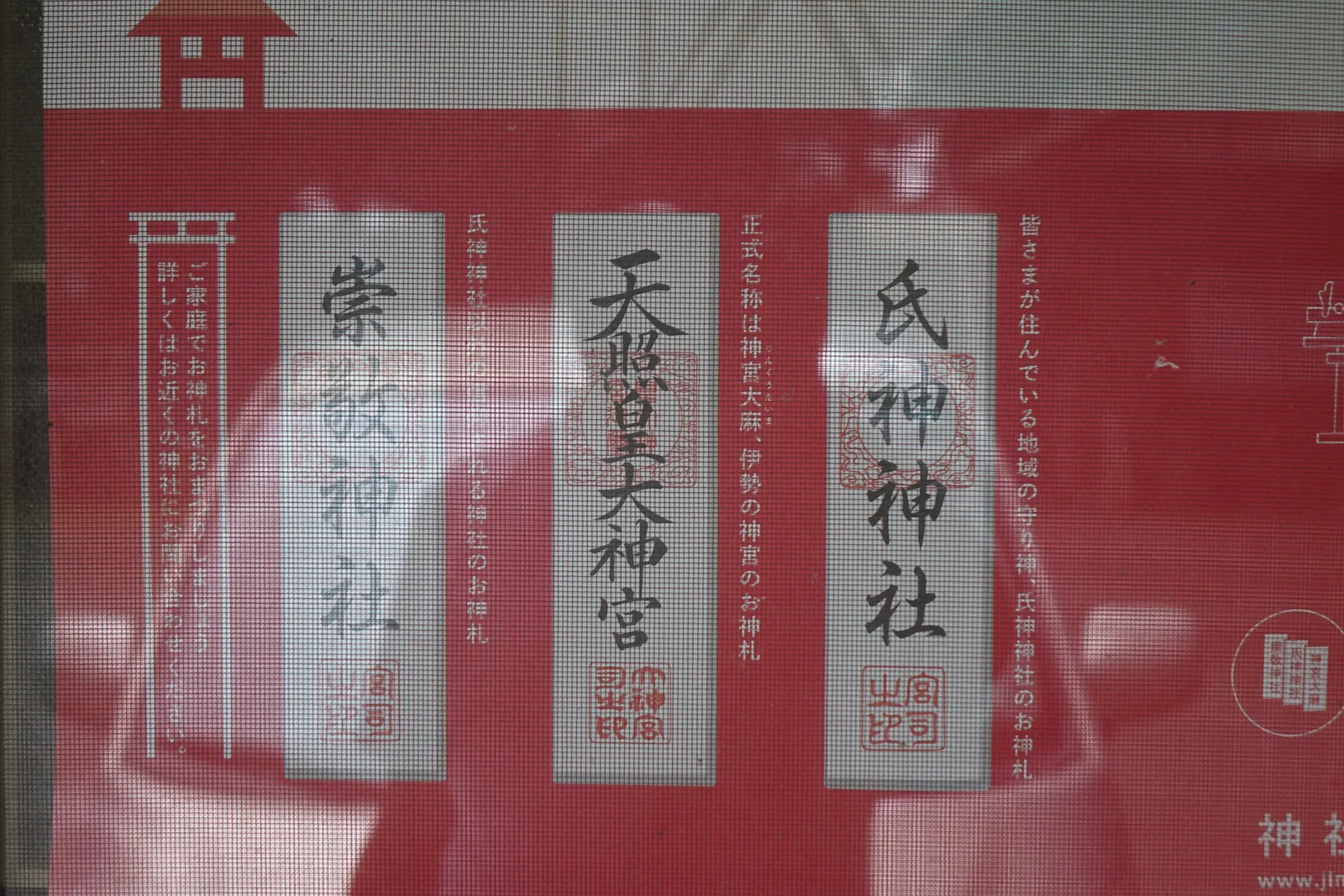 一張紅底的海報，最上緣印著鳥居的圖形。畫面中央並排三張長方形的白色神札圖樣，右起「氏神神社」「天照皇大神宮」「崇敬神社」，每張下方各有一枚朱印。左右兩側是直書的日文說明，講家中神棚該供奉哪幾種神札。海報外面隔著一層紗窗，表面有反光。