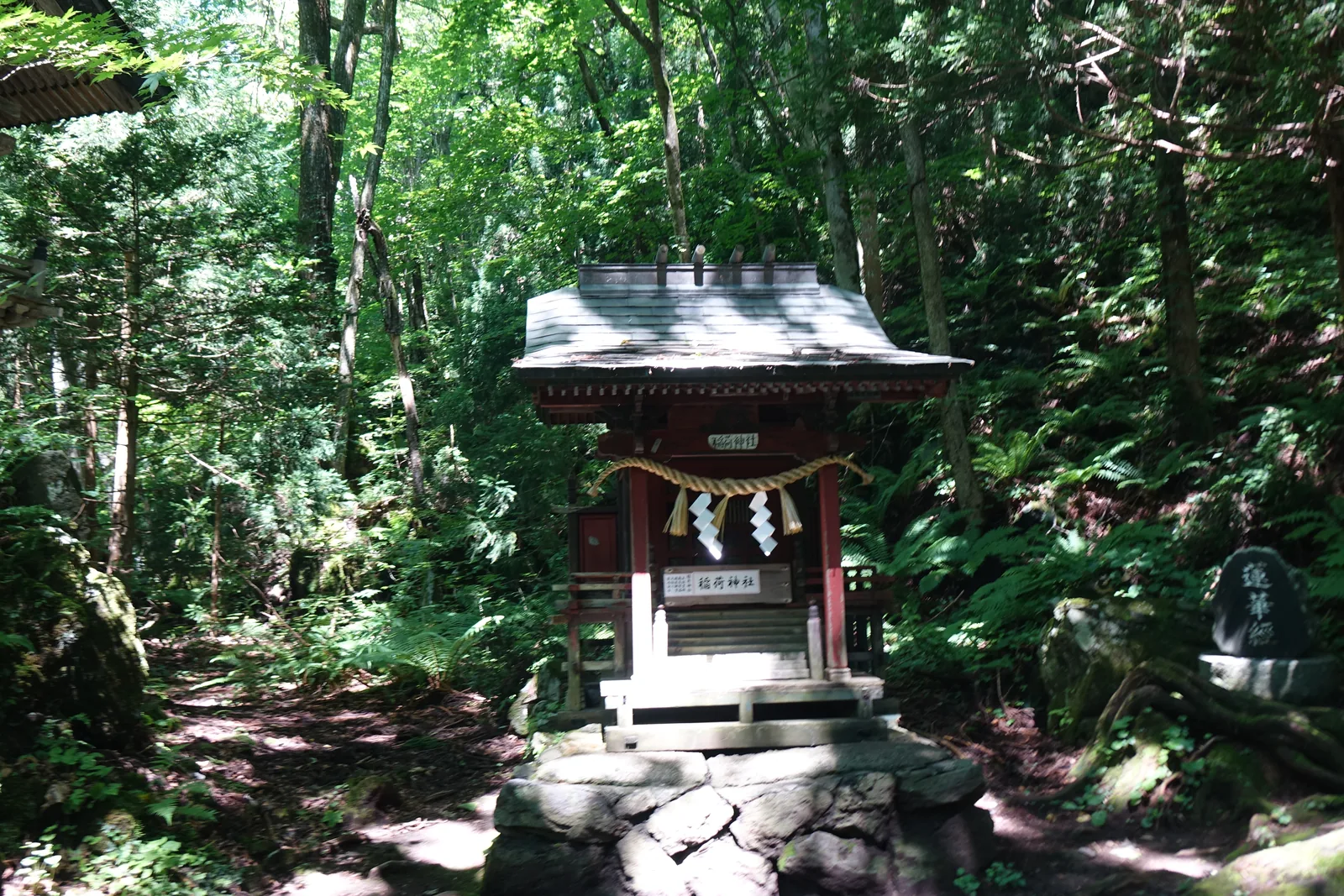一座朱漆的小祠立在一塊岩石砌成的台上，屋面鋪金屬板，簷下懸著扁額，門前橫著一條稻草繩與兩條白紙條，欄杆上另掛一張白紙。扁額與白紙皆書「稲荷神社」。祠的右後方地面上，一塊黑色的石碑刻著「蓮華經」三字。四周是蕨與雜木，光線從樹隙落下。