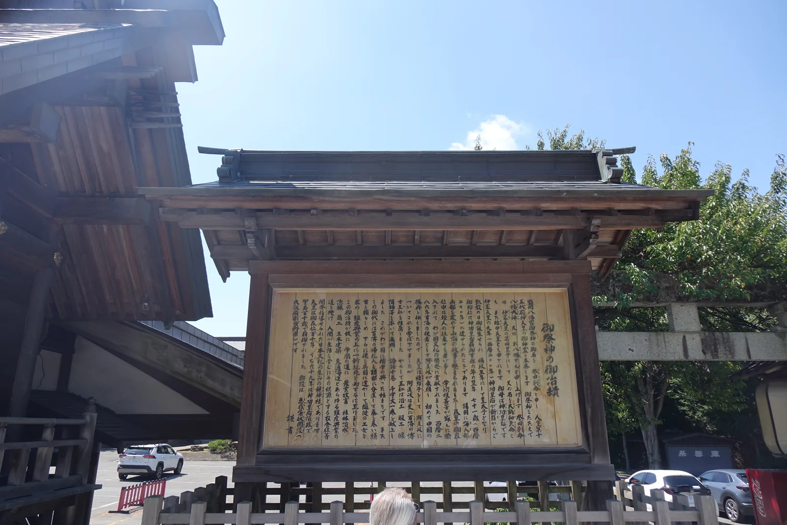 一面銅板葺（どうばんぶき，dōban-buki，銅片鋪成的屋面）屋頂的素木（しらき，shiraki，不上漆的原木）掲示架，框內是一整片直書的木板，標題「御祭神の御治績」。內文從御祭神的名字寫起，一路列到養蠶、農業牧畜、織工、釀造、土木建築、交通海運。板面已經曬成淺褐色，字仍然清楚。背後是社殿的屋簷與停車場。