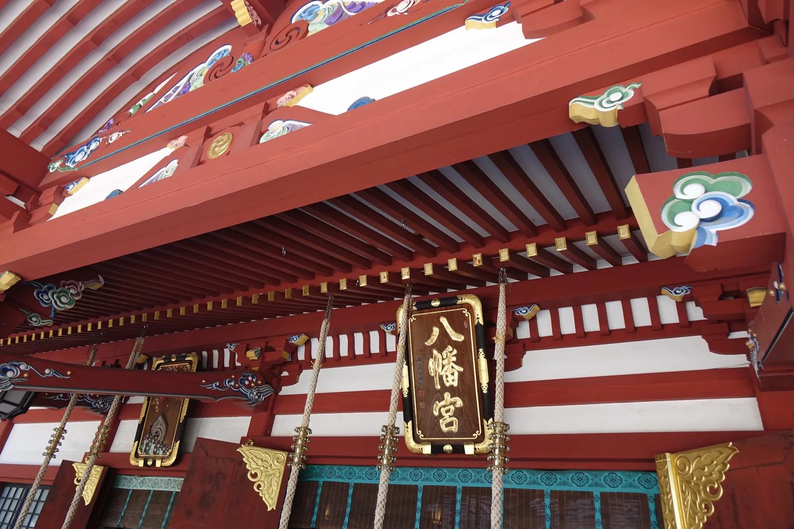 從階前仰看拜殿正面。朱漆的柱與白壁之間，斗栱塗成綠、青、金三色，簷下垂著兩盞金屬燈籠。正中懸著金框的扁額，直書「八幡宮」。左右的橫木上是彩色的雕刻。