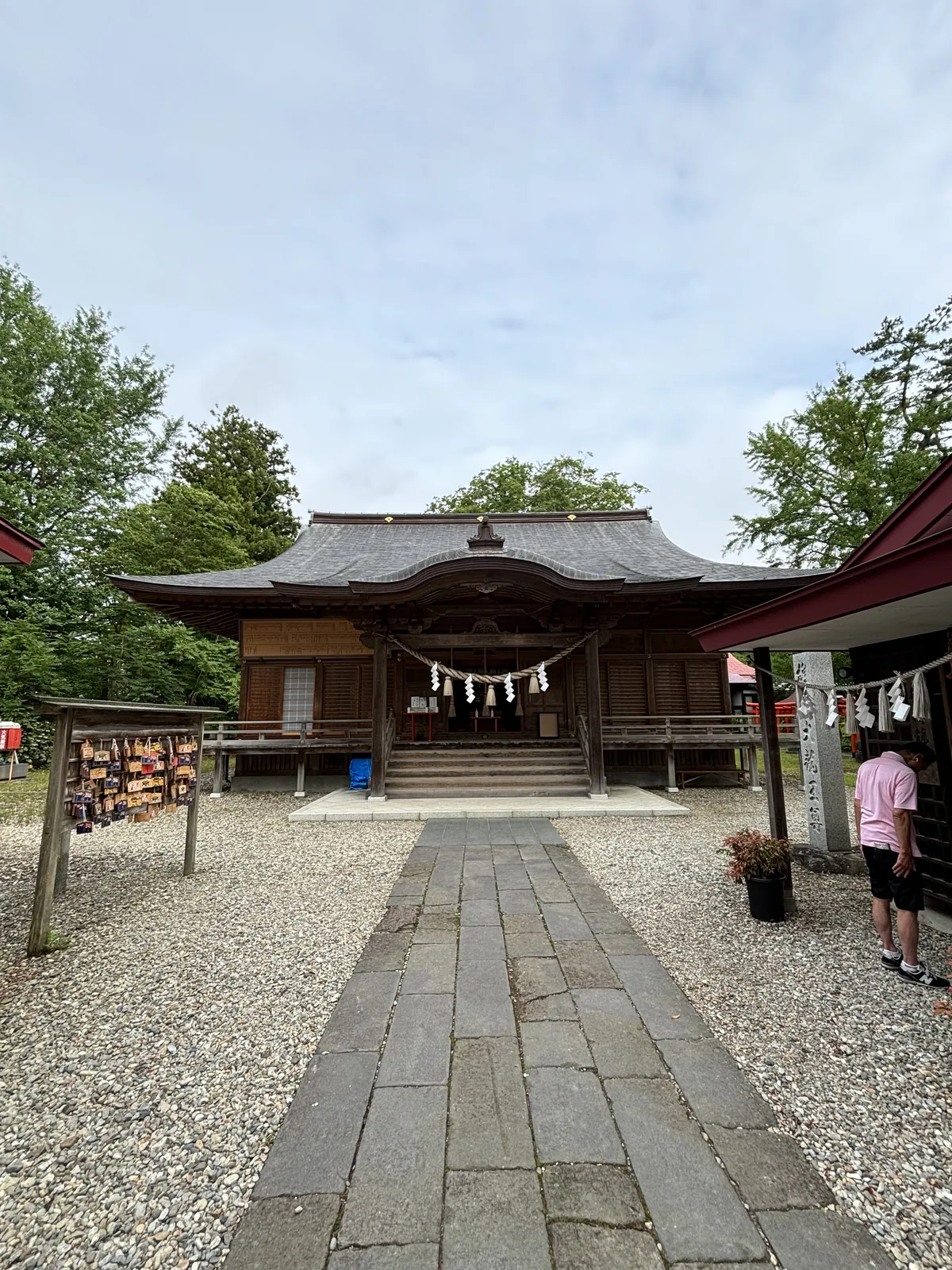 社殿正面。素木（しらき，shiraki，不上漆的原木）的柱與板壁，屋面鋪灰色的金屬板，正中挑出一道中央隆起、兩端下彎的唐破風（からはふ，kara-hafu）。簷下橫著稻草繩，垂下白色的紙垂（しで，shide，摺成階梯狀的白紙）。石板參道從畫面下緣筆直通到階前，兩側鋪白色的玉砂利，左邊是木造的繪馬掛，右邊是朱漆的小建物。天色是陰的。