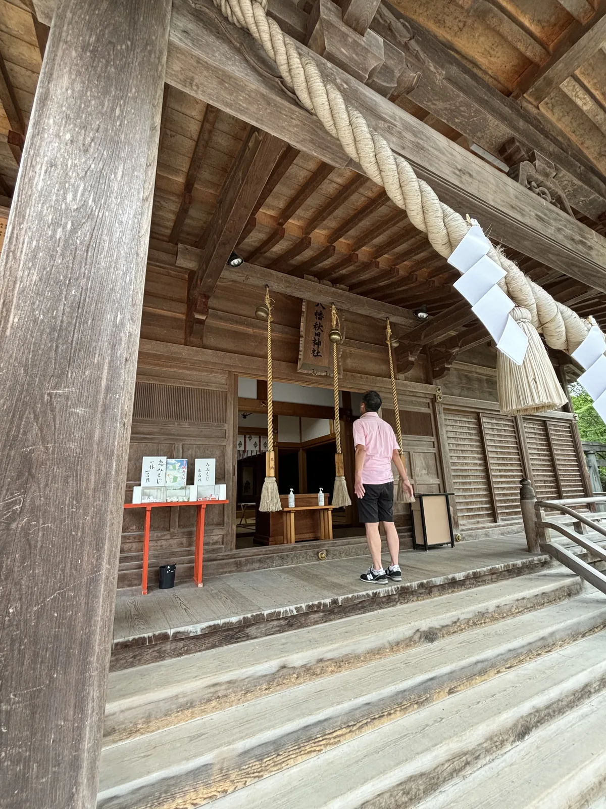 從階下仰看拜殿的開口。粗大的注連繩（しめなわ，shimenawa，標出神域界線的稻草繩）橫過整個正面，垂下四條紙垂（しで，shide，繩上垂下的鋸齒狀白紙）與三條末端結成穗狀的白色鈴緒。深處是木格門與一塊金框的小扁額。階前擺著一張鋪紅布的長桌，桌上是幾疊印刷品；一個穿粉紅上衣的人站在階上，背對鏡頭。