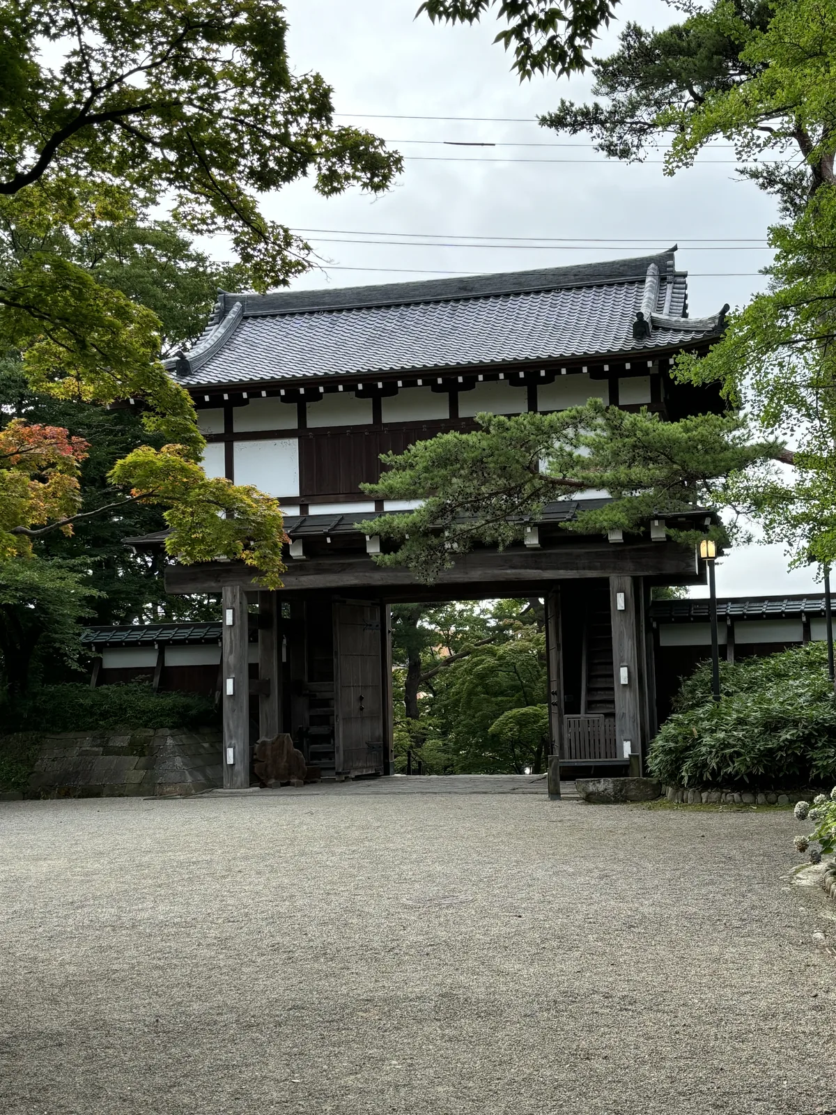 木造二層、瓦葺的櫓門立在石階之上，門扇是厚重的板門，釘著成排的金屬鋲。門的兩側是白灰牆與木板圍牆，前方是平整的砂礫地與修剪過的樹。