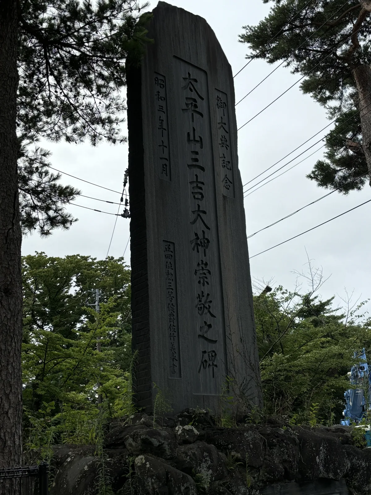 一塊高而窄的深色石碑立在樹叢之間，正面直書「太平山三吉大神崇敬之碑」，右側小字「御大典記念」，左側是「昭和三年十一月」。碑的上方是松枝與電線，下方是石砌的基座與草。
