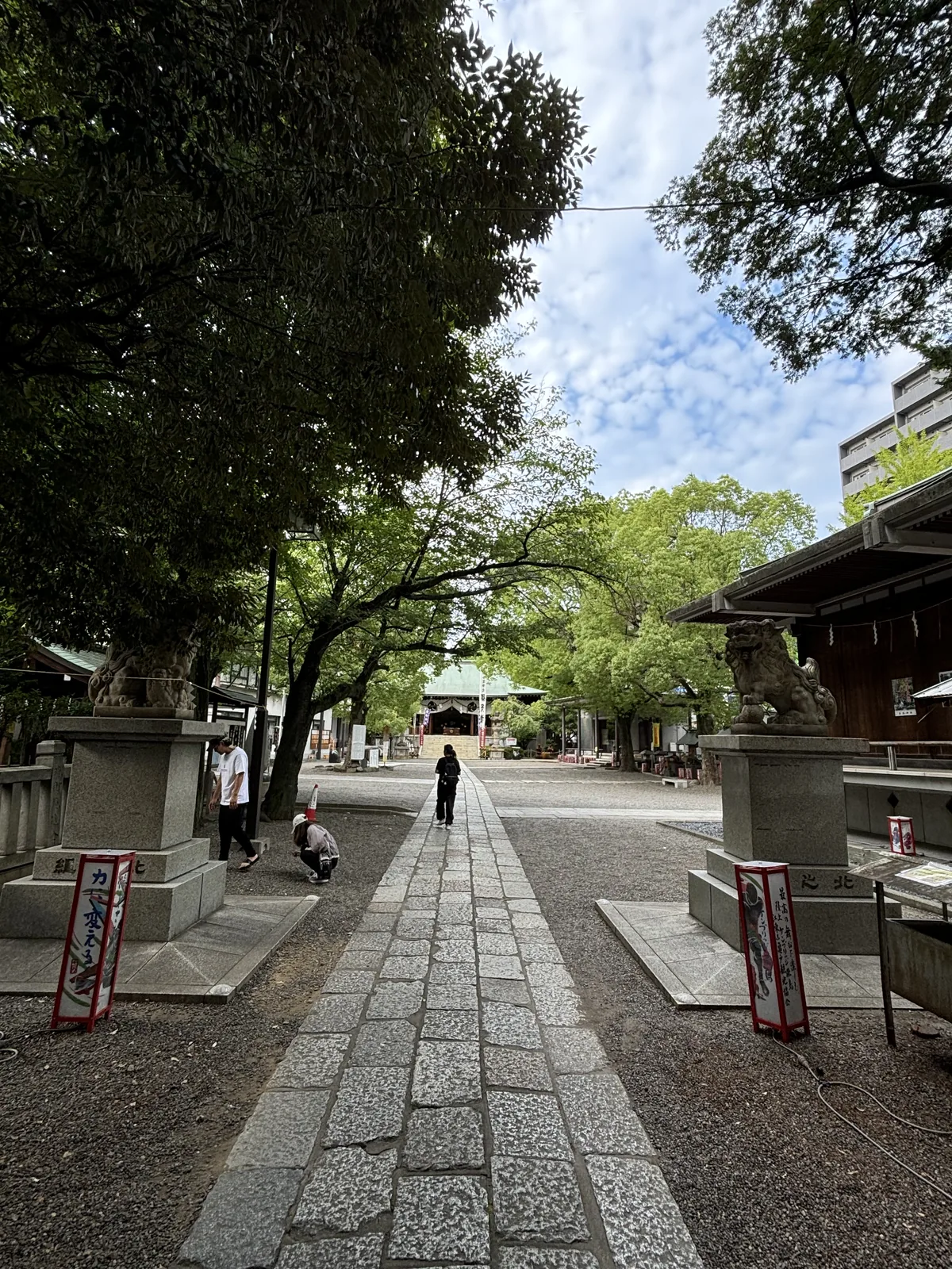 從鳥居下往內看。石板鋪成的直線參道從畫面下緣延伸到深處的社殿，兩側是低矮的境內建物、樹與石燈籠。左前方立著一面紅色的幟旗。一個人影走在參道盡頭。地面反著陰天的光。