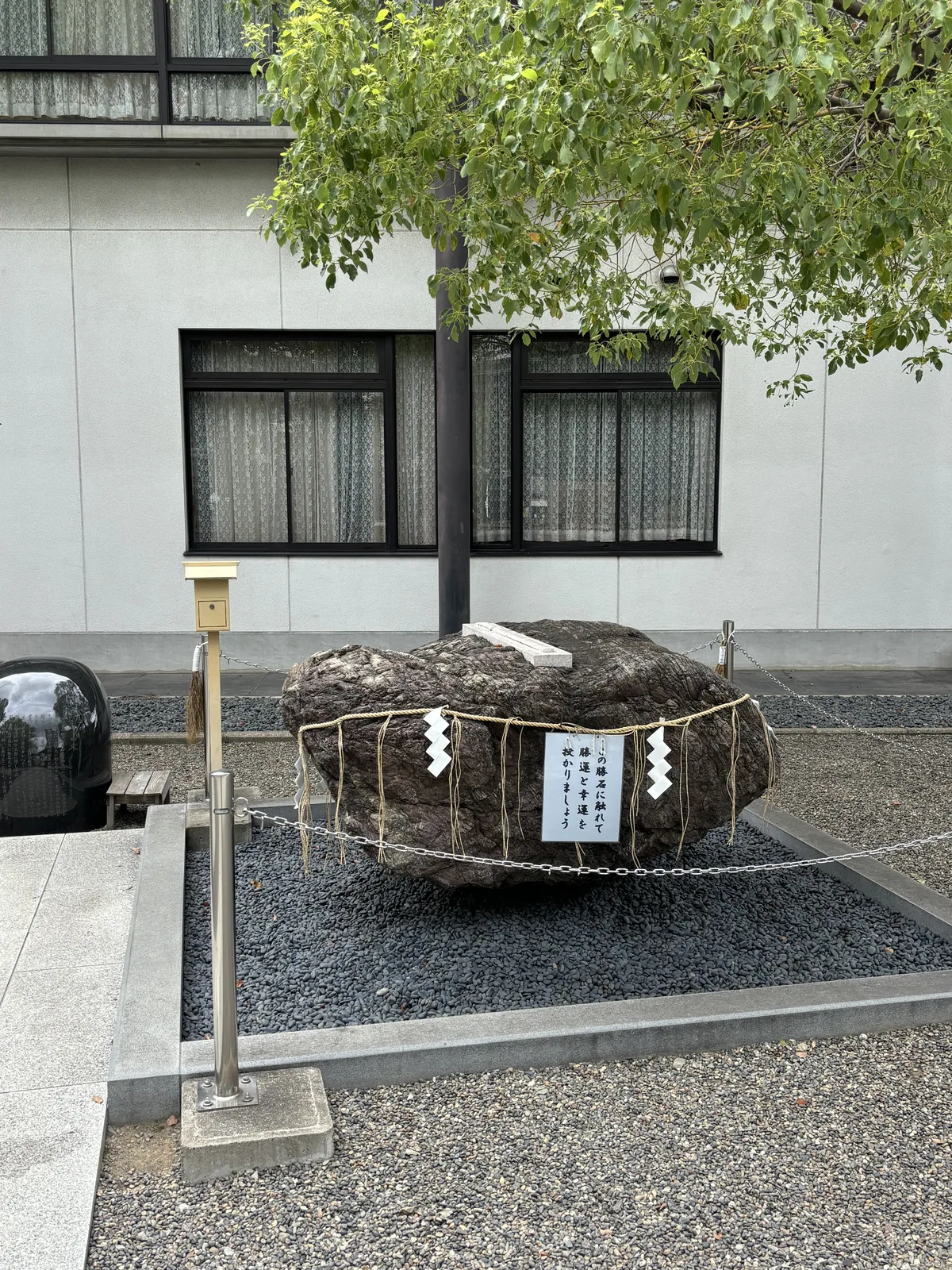 一塊臥倒的褐色大石放在黑色礫石鋪成的方框裡，四角立著金屬柱、拉著鐵鏈圍起來。石腰繫一條稻草繩，垂下白色的紙垂（しで，shide，摺成階梯狀垂在繩上的白紙）。石頂平放一塊白色的石雕，形狀是一把太刀。石前掛著一張白牌，寫「勝石に触れて　勝運と幸運を授かりましょう」。背後是鄰棟住宅的窗。